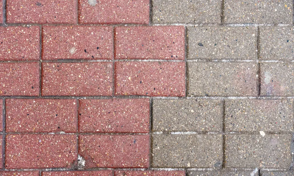 A close-up, overhead view of a brick-paved surface divided into two distinct sections. The left side features red bricks with small flecks of white and black, appearing somewhat wet and showing signs of light frost or dirt along the grout lines. The right side is composed of gray concrete pavers, also with visible flecks and a similar pattern of frost or dirt along the grout lines. The image appears to be a comparison of a section of pavement before and after cleaning, as suggested by the alt text.
