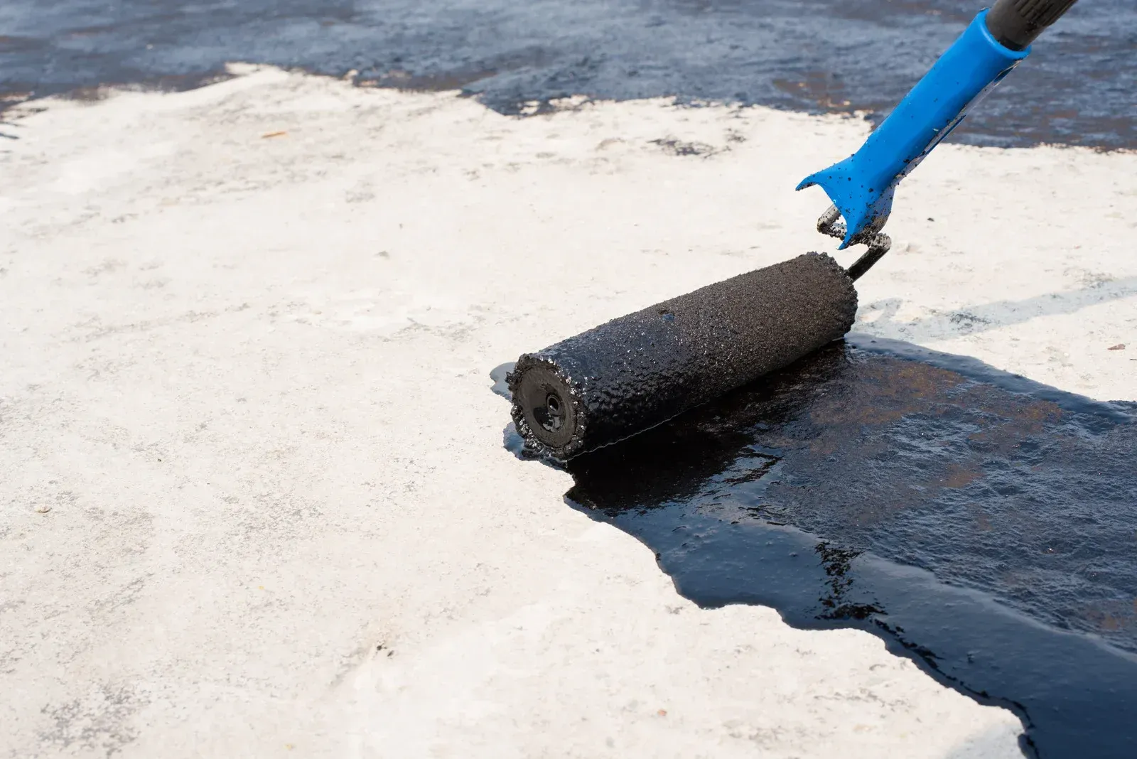 A close-up of a blue-handled roller applying a thick, black sealant or coating onto a light-colored, possibly concrete, surface.