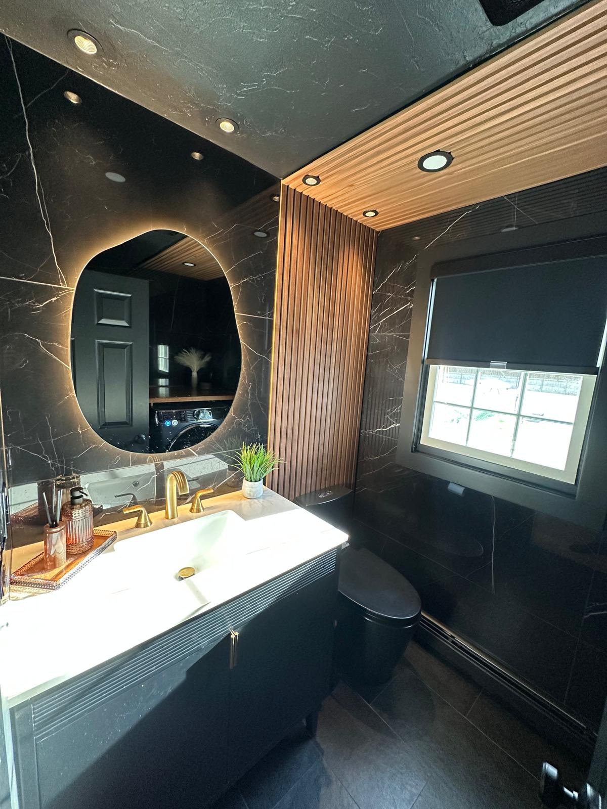 Complete bathroom renovation featuring a modern luxury design. This project included the installation of a custom floating vanity, contemporary vessel sink, illuminated oval mirror, vertical wood accent wall panels, updated plumbing fixtures, and premium finishes throughout. The dark color palette combined with warm wood tones creates an elegant and sophisticated atmosphere.