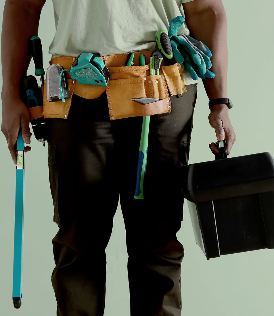 A person is wearing a tool belt loaded with various tools and is holding a toolbox and a level.