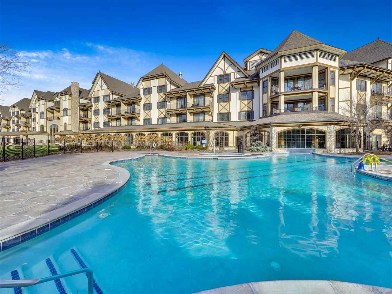 A large, blue swimming pool with clear water is in the foreground, reflecting the bright blue sky. Beyond the pool, a multi-story building with a Tudor-style architectural design stands tall. The building features numerous balconies, windows, and a distinctive peaked roofline. Trees with bare branches are visible on the sides of the building, suggesting a cooler season. The overall scene depicts a resort or hotel complex with outdoor amenities.