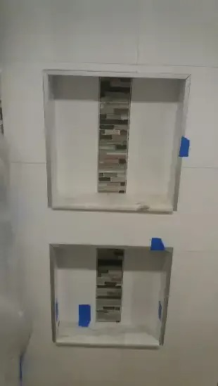 Two rectangular niches with mosaic tile backsplashes are set into a white tiled shower wall. Blue painter's tape outlines the niches, indicating recent construction or finishing work.