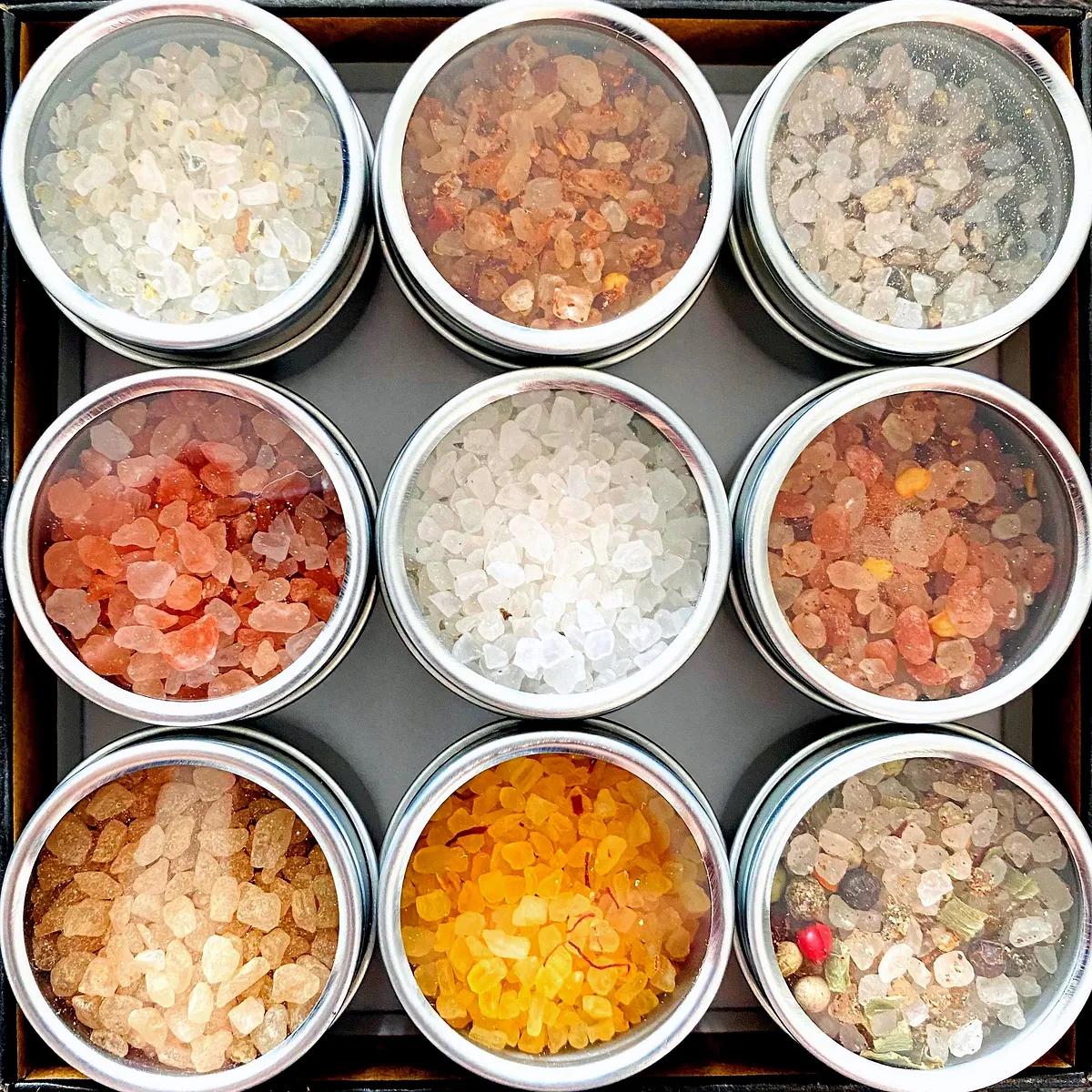 A top-down view of nine small, circular metal tins filled with various types of salt and spices. The tins are arranged in a 3x3 grid within a dark box. The salts and spices vary in color, including white, pink, orange, brown, and mixed tones with visible herbs and peppercorns. Some tins have clear lids, allowing the contents to be seen. The lighting highlights the crystalline textures of the salts and the vibrant colors of the spices.