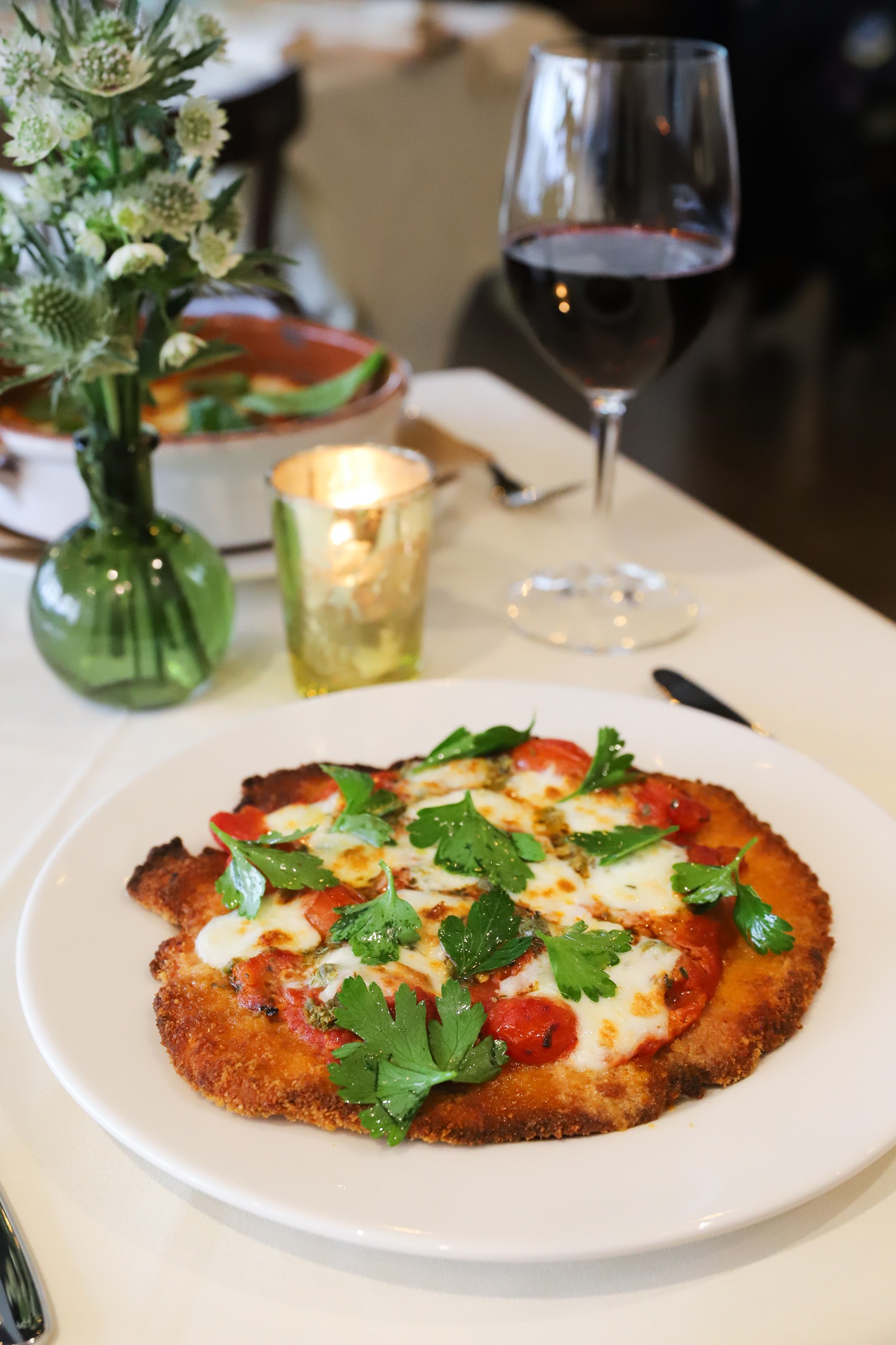 A breaded chicken cutlet topped with marinara, melted cheese, and fresh herbs served on a white plate next to a glass of red wine and a candle.