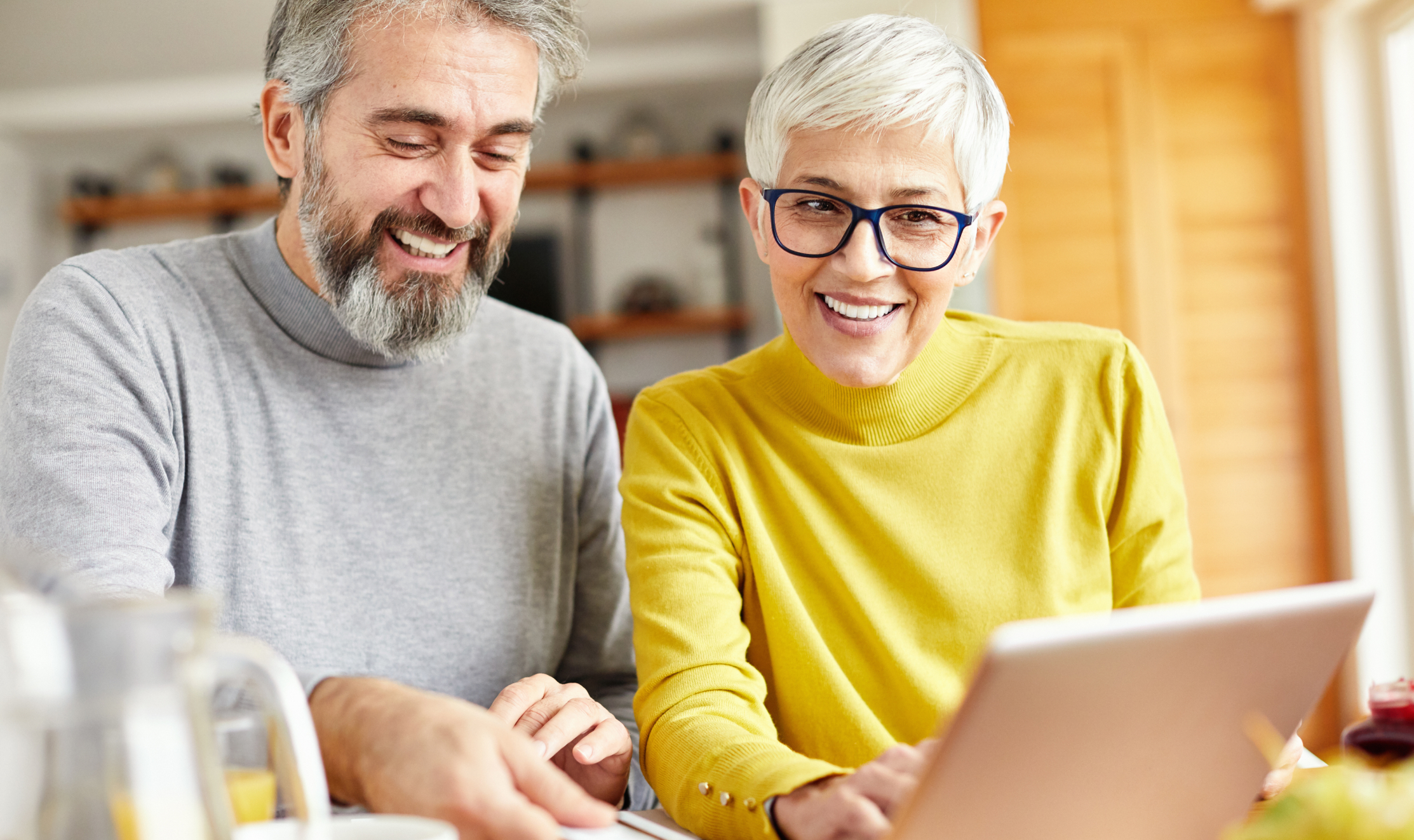 A smiling older couple, a man with a gray beard and a woman with short white hair and glasses, are looking at a laptop on a table. The man is wearing a gray turtleneck, and the woman is wearing a yellow turtleneck. They appear to be in a home setting, possibly a kitchen or dining room, with a pitcher of orange juice and a jar of jam visible on the table.