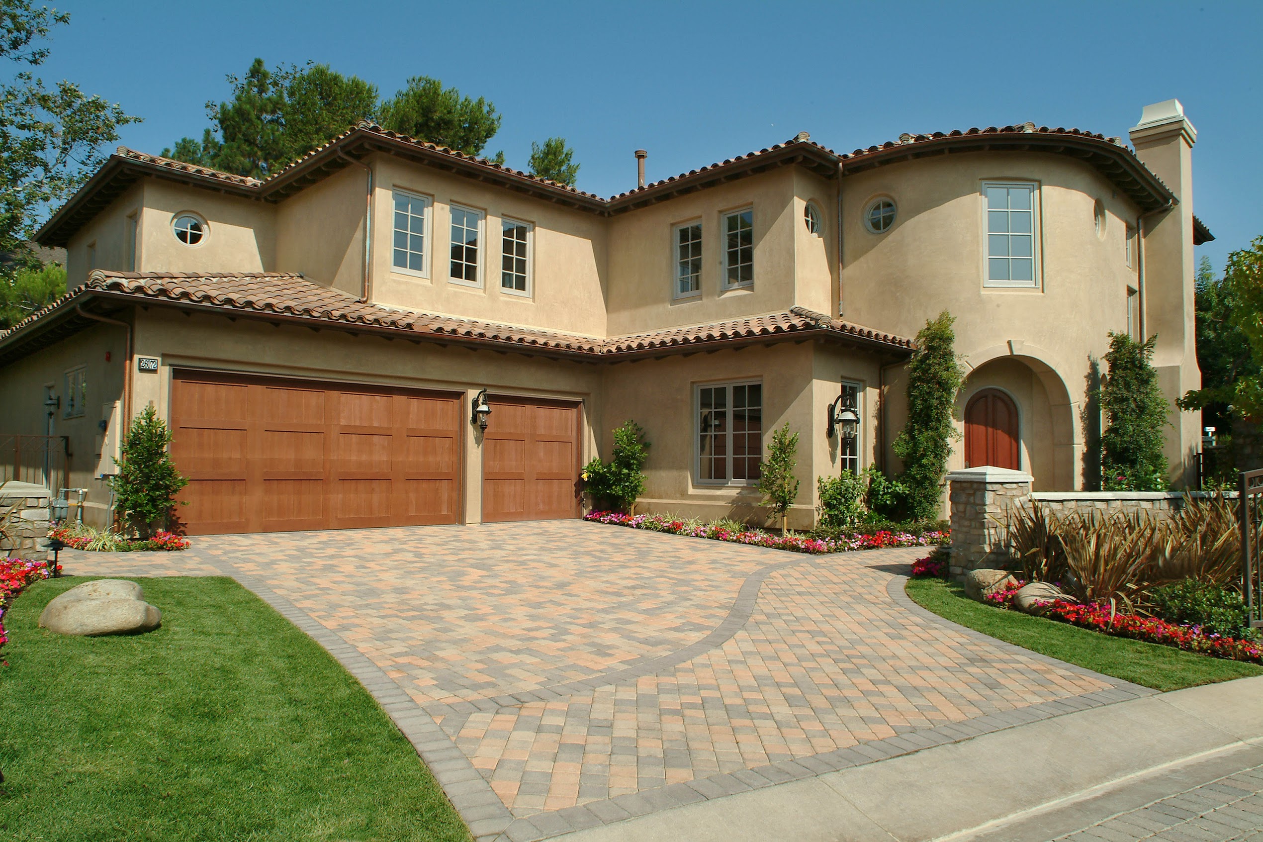 Elegant Spanish-style stucco mansion with tiled roof, arched entry, wooden garage, landscaped driveway, flower beds, and manicured lawn.