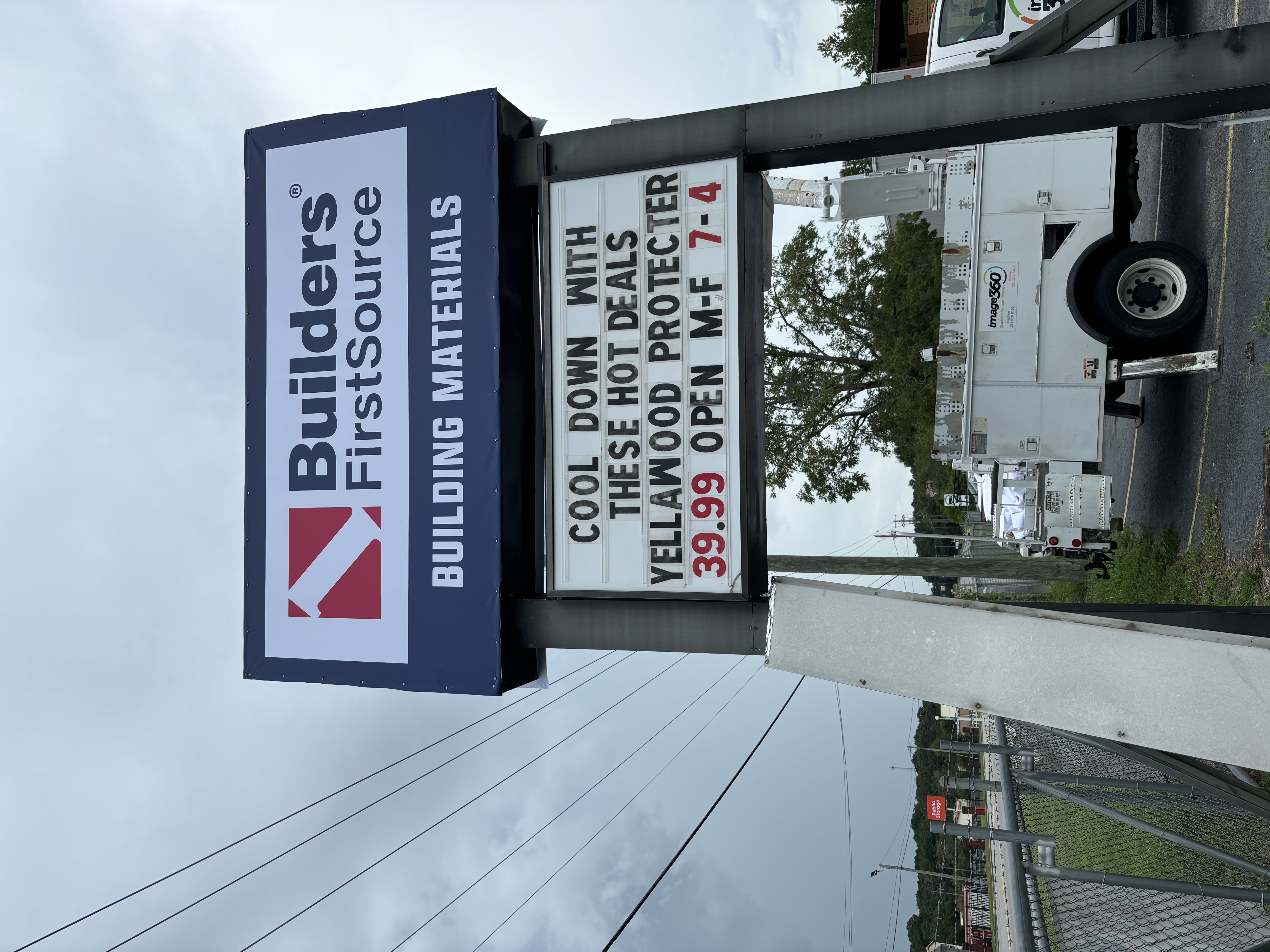 A tall Builders FirstSource street sign with the message 'Cool Down With These Hot Deals - YellaWood Protector $39.99 - Open M-F 7-4.' The sign is positioned alongside a road with utility vehicles and a chain-link fence visible nearby under an overcast sky.