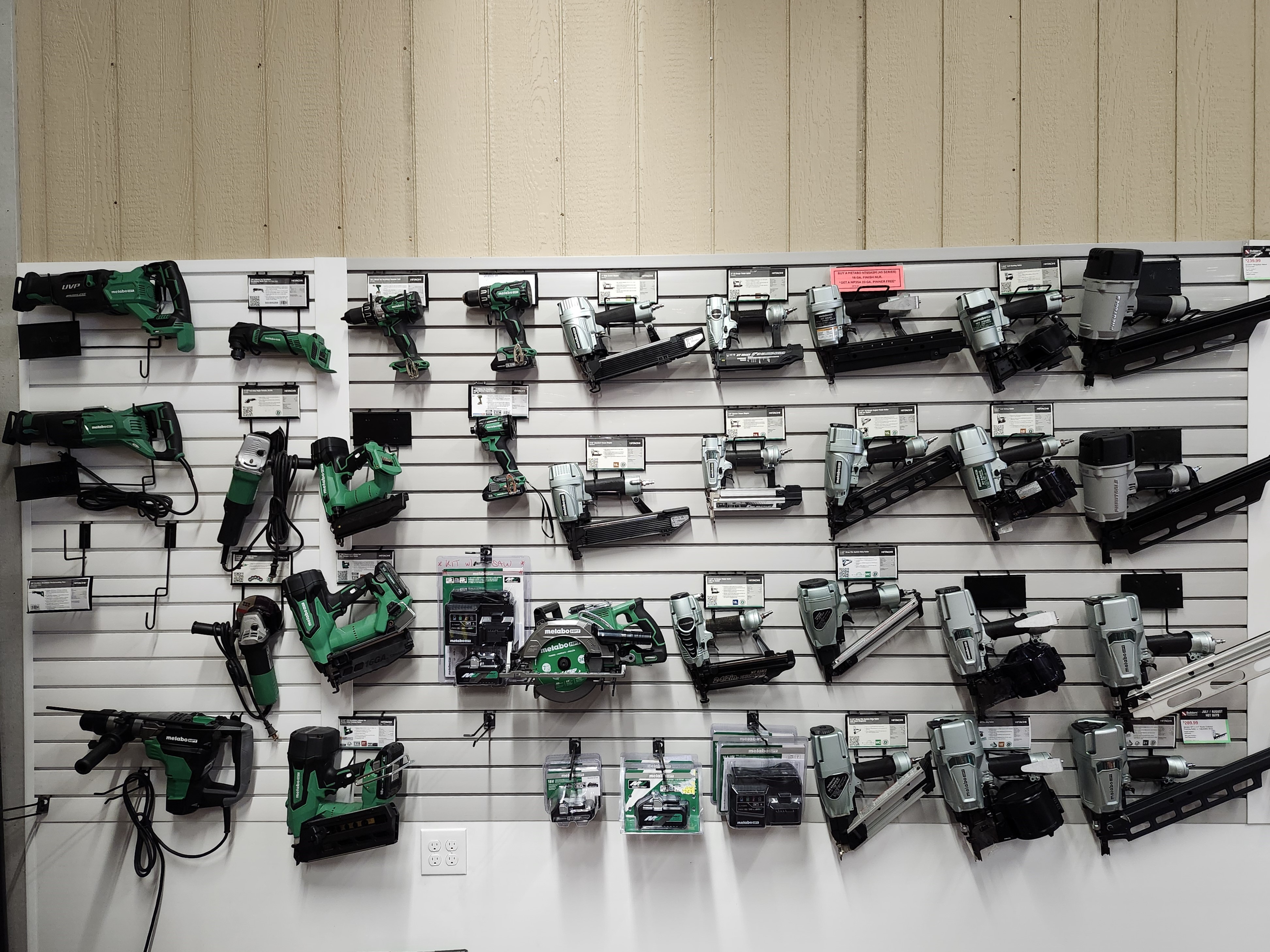 An extensive display of Metabo power tools and nailers at a Builders FirstSource lumber yard. The display includes various models of nail guns, drills, and saws, all arranged on a slat wall. The tools are displayed with product labels and information, emphasizing the Metabo brand.