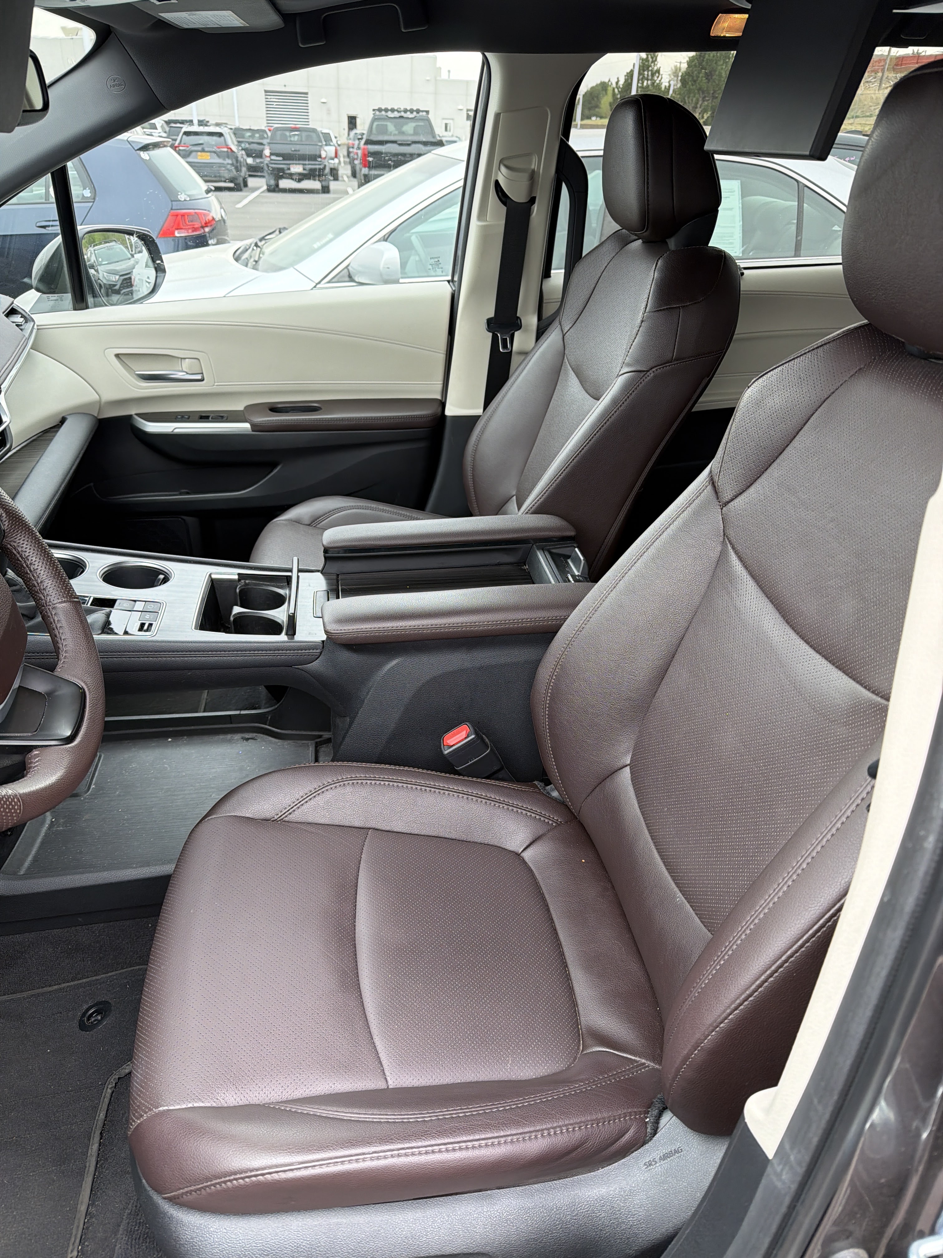 Interior view of a car, showing the front passenger seat and part of the driver's seat and center console. The seats are upholstered in perforated brown leather with cream-colored accents on the door panels. The dashboard features wood-grain trim. In the background, through the windows, several other cars are parked in a lot.