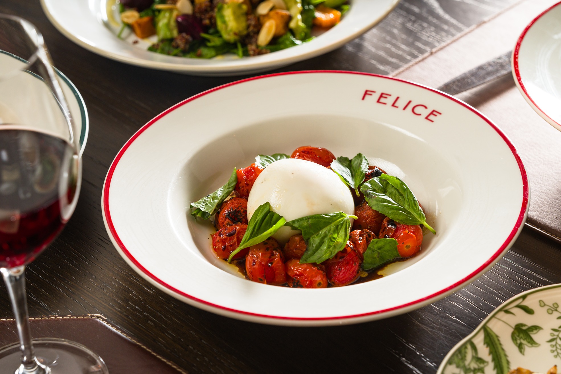 A white Felice-branded bowl holds a ball of fresh burrata surrounded by roasted cherry tomatoes and basil. A glass of red wine and a salad are partially visible on the dark wood table.