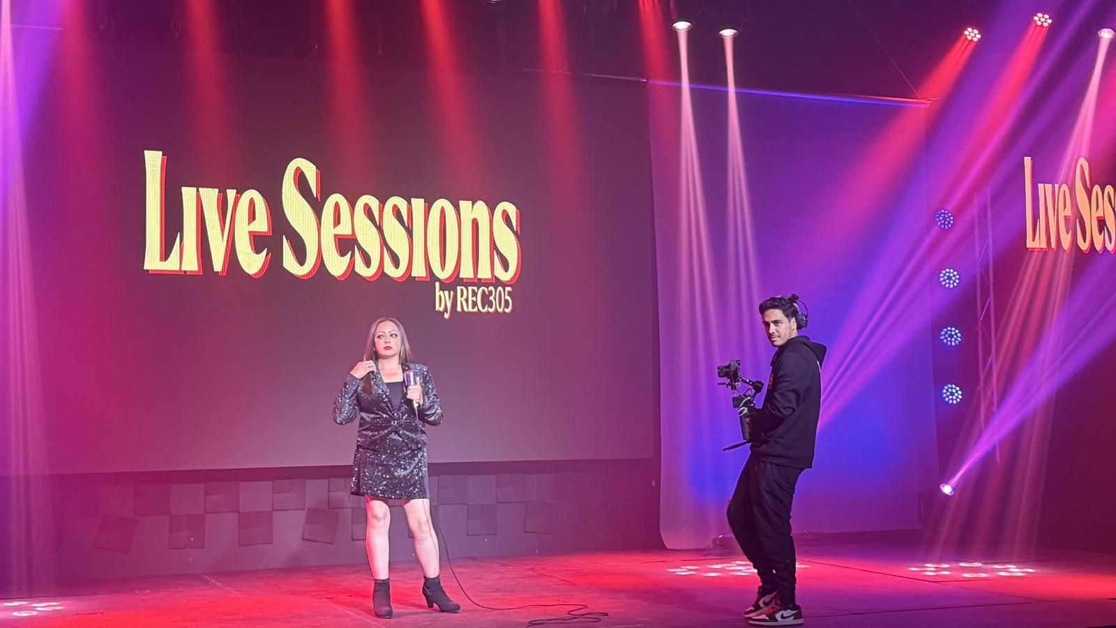 En esta imagen te mostramos la versatilidad de nuestro espacio configurado como un estudio de televisi&oacute;n. En el centro del escenario se encuentra una cantante solista, mientras un camar&oacute;grafo de nuestro equipo, equipado con un estabilizador electr&oacute;nico, se mueve para registrar sus movimientos con calidad profesional. La escena est&aacute; ambientada con luces de fondo p&uacute;rpuras y rojas. Si buscas salones para conferencias o eventos para presentaciones con transmisiones digitales de alta definici&oacute;n, somos el sal&oacute;n para eventos indicado.