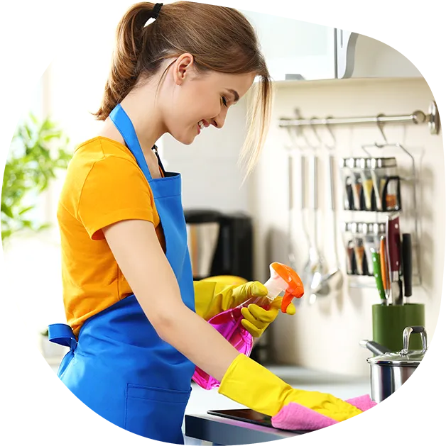 A woman wearing an orange t-shirt, a blue apron, and yellow gloves is cleaning a kitchen counter. She is holding a pink spray bottle with an orange nozzle in her right hand and a pink cleaning cloth in her left hand. She is smiling and looking down at her work. The background shows a kitchen with a rack of spices and utensils, and a window with greenery outside.