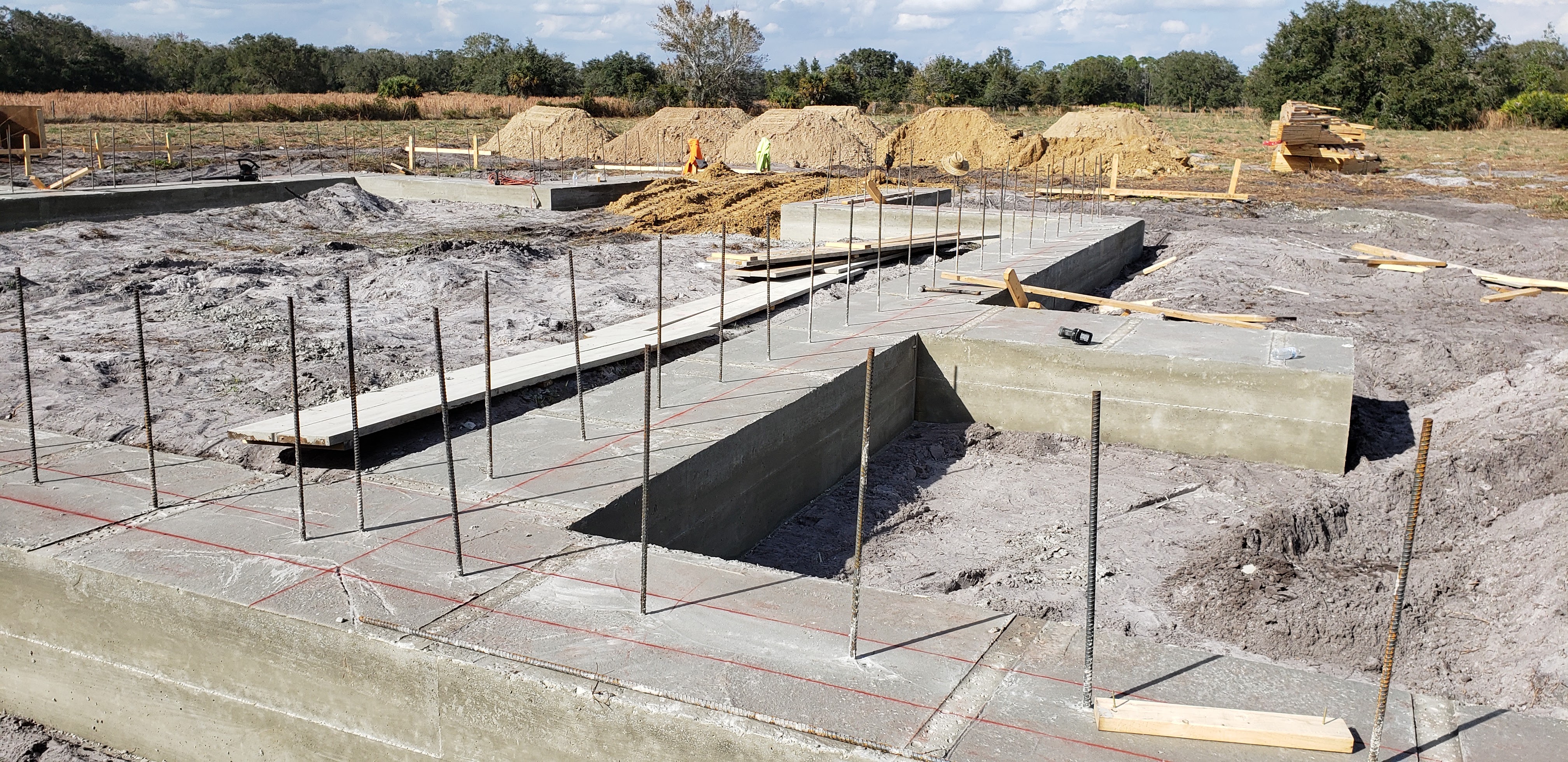 A construction site for a house foundation is shown, with concrete footings and rebar in place. Pile 