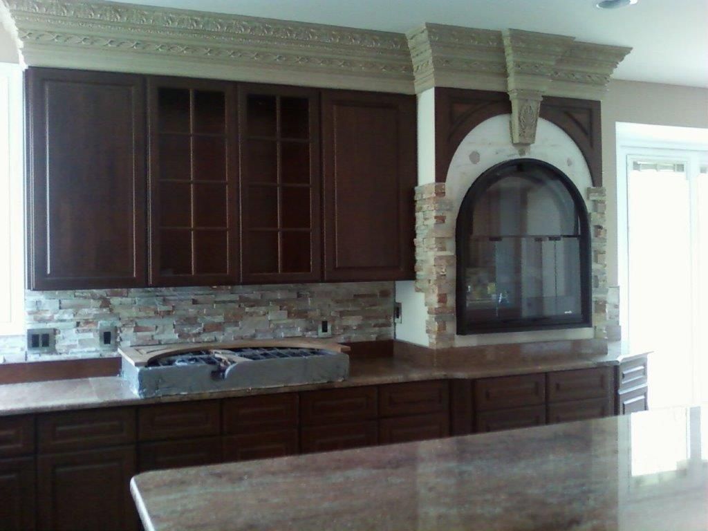A custom kitchen design featuring dark wooden cabinetry, stone backsplash, and ornate crown molding. An arched niche with decorative stonework adds a focal point above the countertop workspace area.