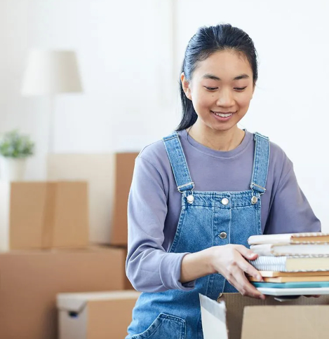 Young woman in denim overalls carefully packing a stack of books into a cardboard moving box surrounded by packed boxes.