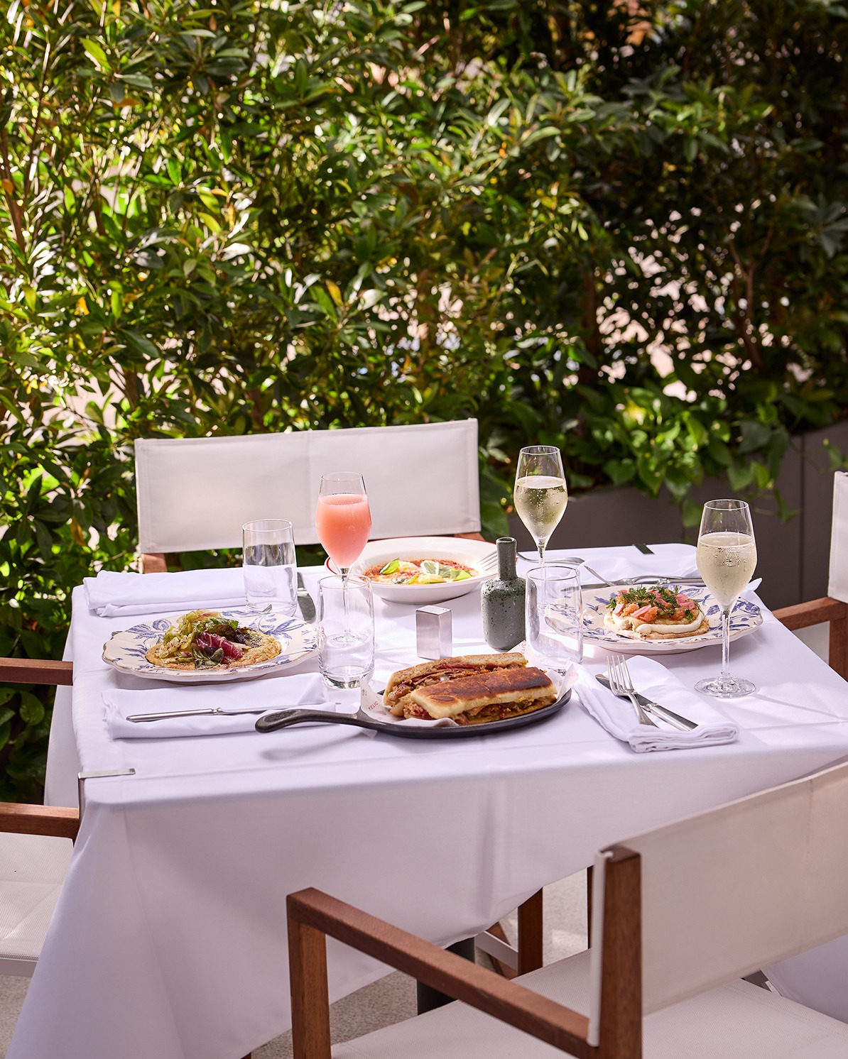A sun-drenched outdoor table featuring a panini, seafood appetizers, and cocktails, surrounded by lush green privacy hedges.