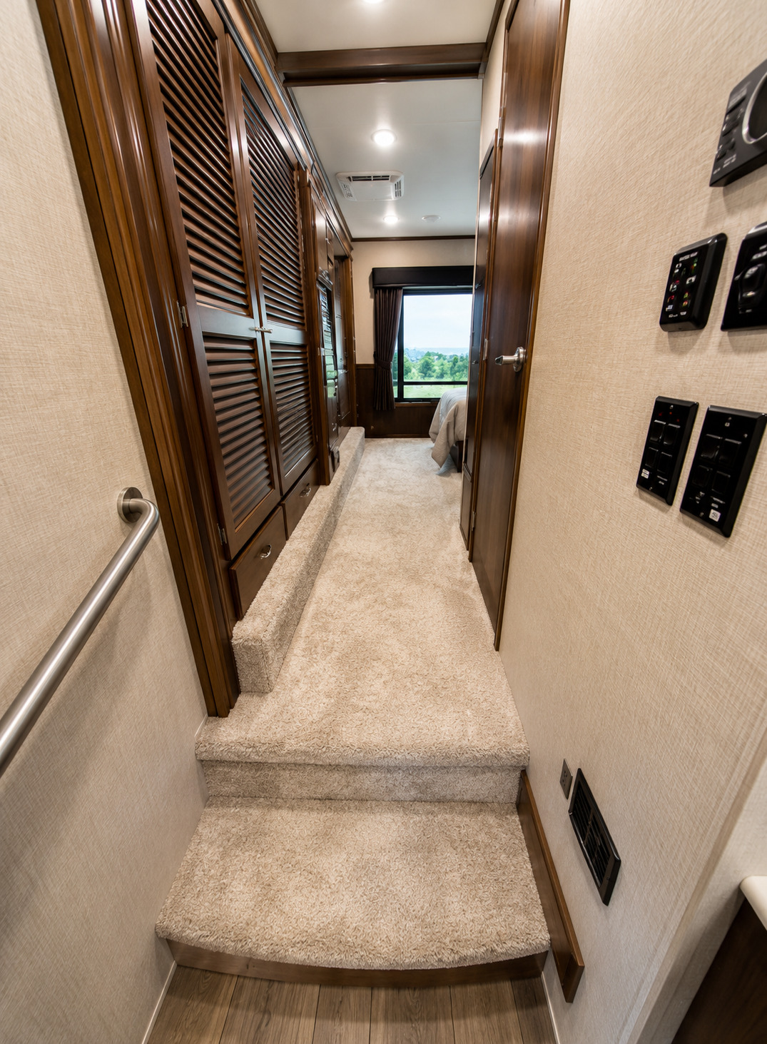 Luxury RV interior hallway with carpeted steps, dark wood cabinetry, louvered closet doors, wall-mounted control panels, recessed lighting, and a bedroom visible ahead through an open doorway with window views.