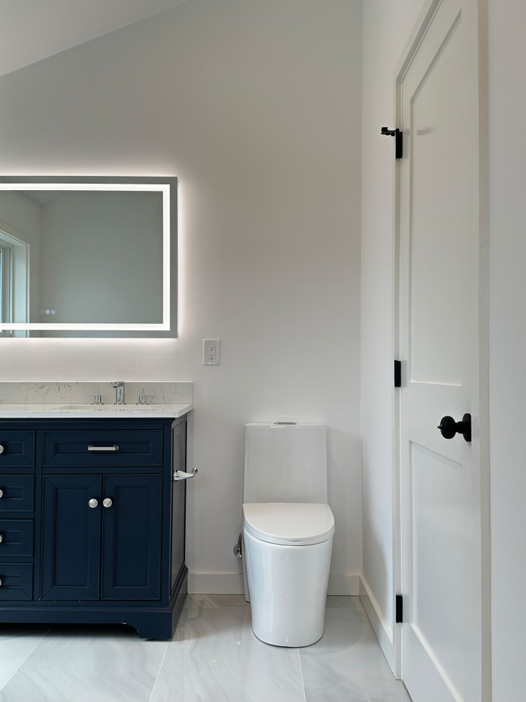 A modern bathroom featuring a navy blue vanity with a white marble countertop, a white toilet, and a large illuminated mirror. A white door with black hardware is visible on the right side.