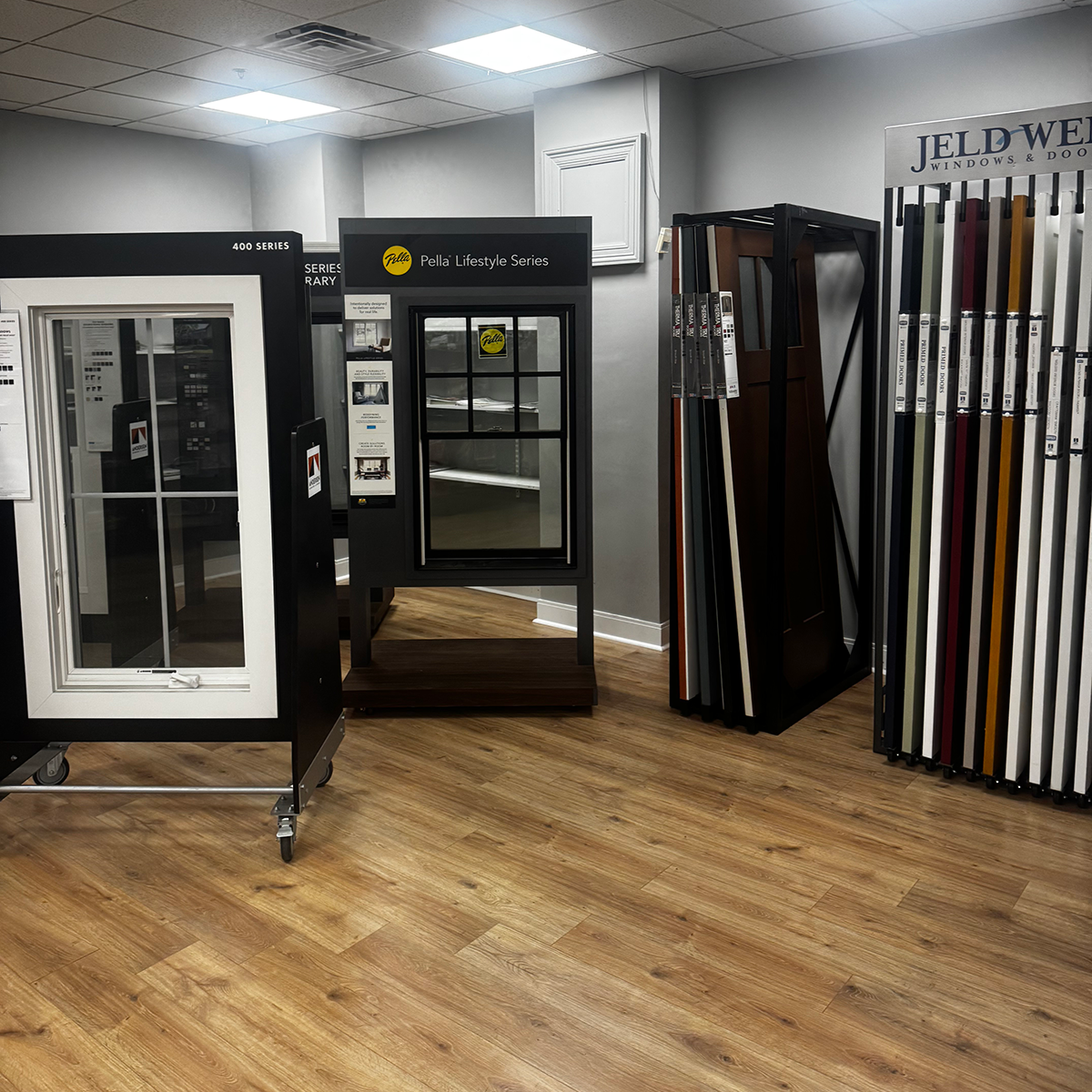 Interior showroom display featuring window and door samples on stands and racks. A black-framed window with divided panes is labeled “Pella Lifestyle Series” in the center, while a white window display and multiple full-height door samples are arranged nearby. Color swatches, product information and a JELD-WEN Windows & Doors sign are visible against gray walls and a light wood-look floor.