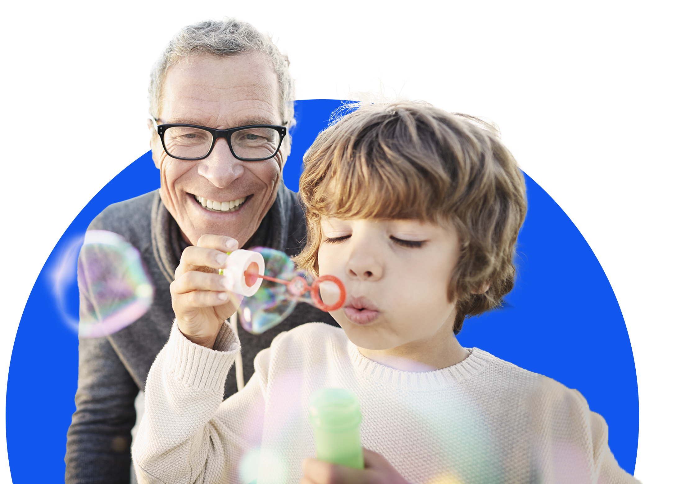 A man wearing glasses and a gray sweater is smiling while standing behind a young boy who is blowing bubbles. The boy is wearing a cream-colored sweater and is focused on blowing bubbles from a wand connected to a small green bottle.