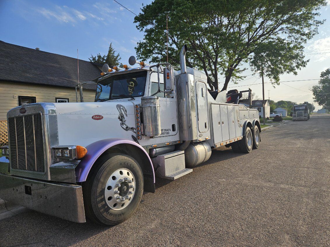 A large white Peterbilt heavy-duty tow truck with purple front fender accents is parked along a quiet residential street. The truck features prominent chrome exhaust stacks, grille, and front wheels.