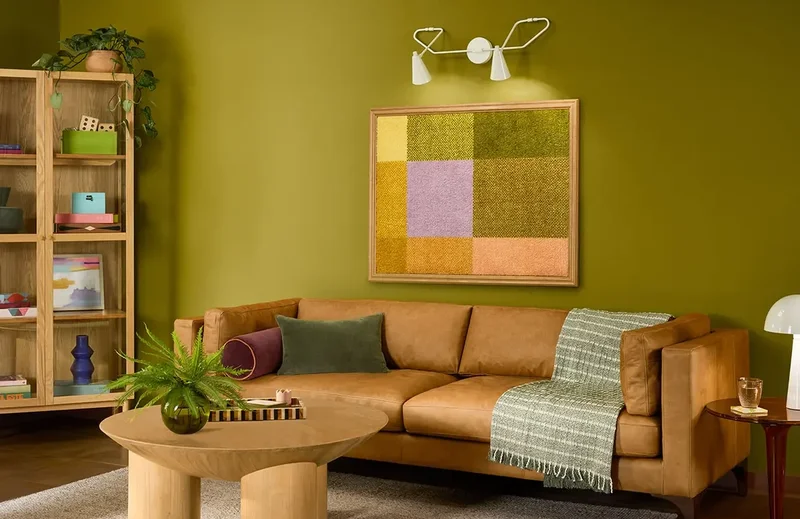A cozy living room with a camel-colored leather sofa, a wooden coffee table, and a large abstract artwork on a green wall. The room is decorated with plants, books, and decorative items, creating a warm and inviting atmosphere.