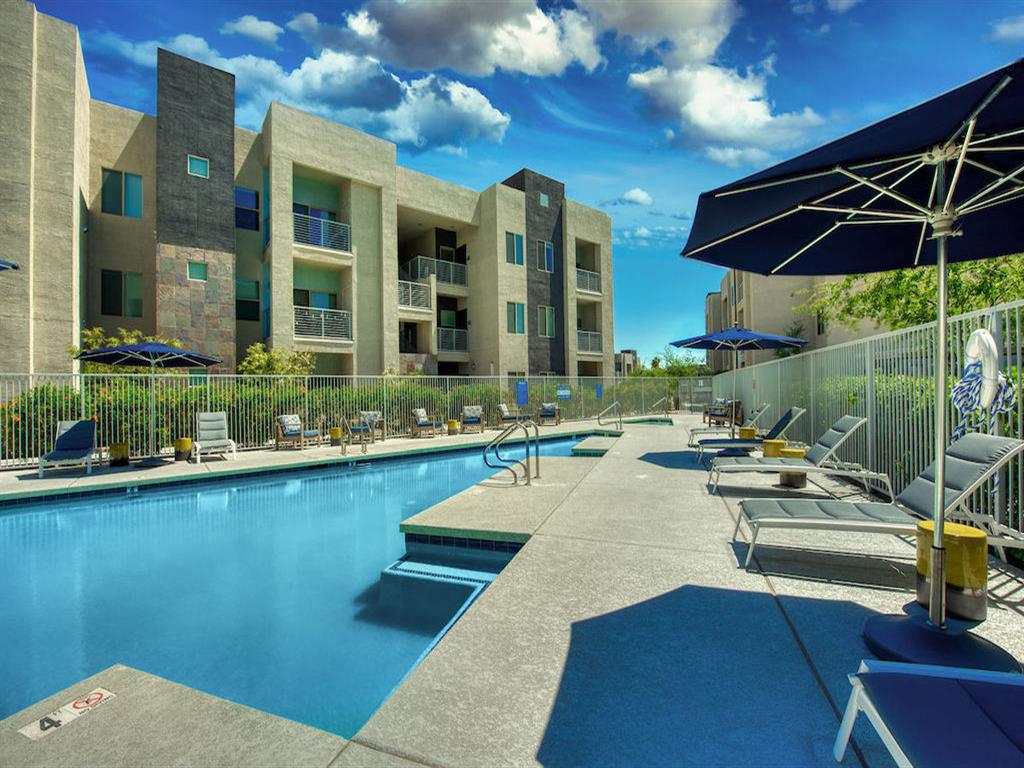 Pool with buildings in the background and patio umbrella up front. Four lounge chairs line the pool.