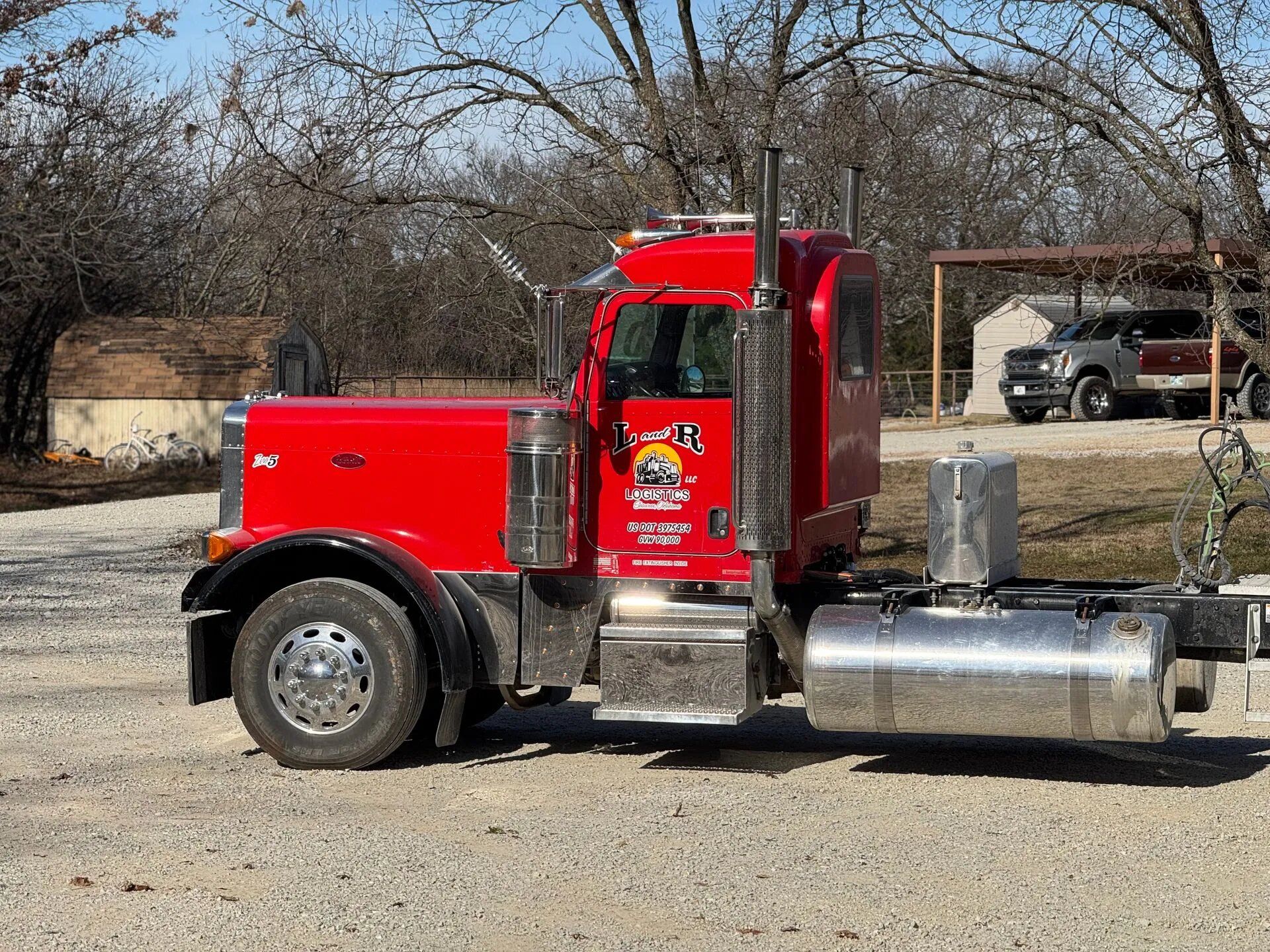 L & R Logistics LLC red semi truck photographed from the side, displaying its cab, chrome fuel tank, exhaust stacks, branded door, and heavy-duty transportation equipment outdoors locally in Oklahoma.