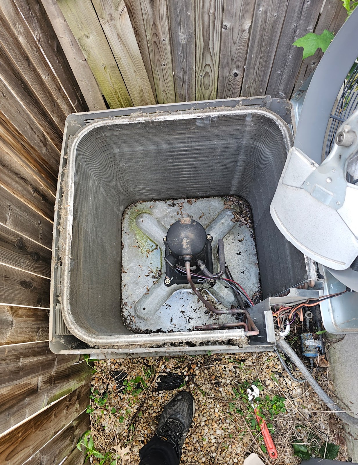 A top-down view of the interior of an air conditioning unit, with its cover removed, revealing the condenser coils, fan motor, and wiring. The unit is situated outdoors, adjacent to a wooden fence, with debris and a person's shoe visible on the ground surrounding it. A red utility knife and some white material are also present on the gravel.