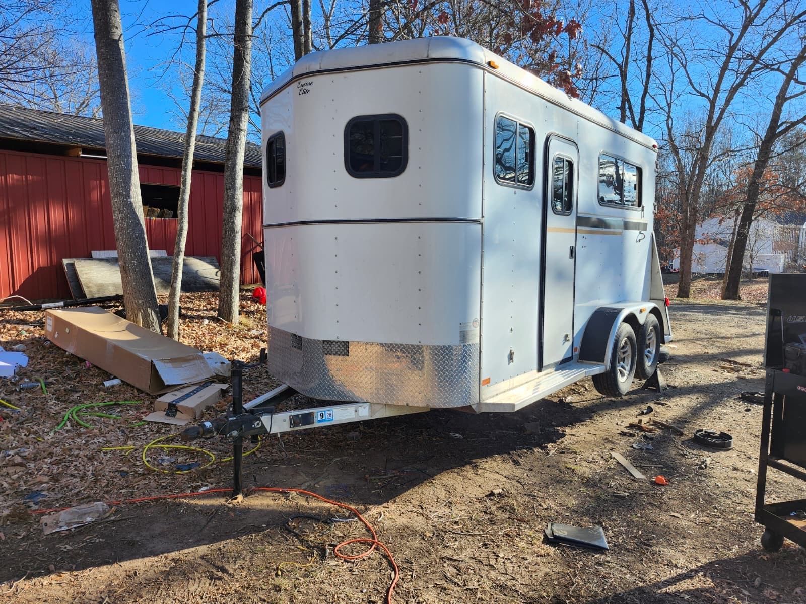 A white enclosed trailer is parked outdoors near a red building and trees. The trailer features dual axles, side door access, and a clean exterior, representing storage or transportation use for equipment or goods.