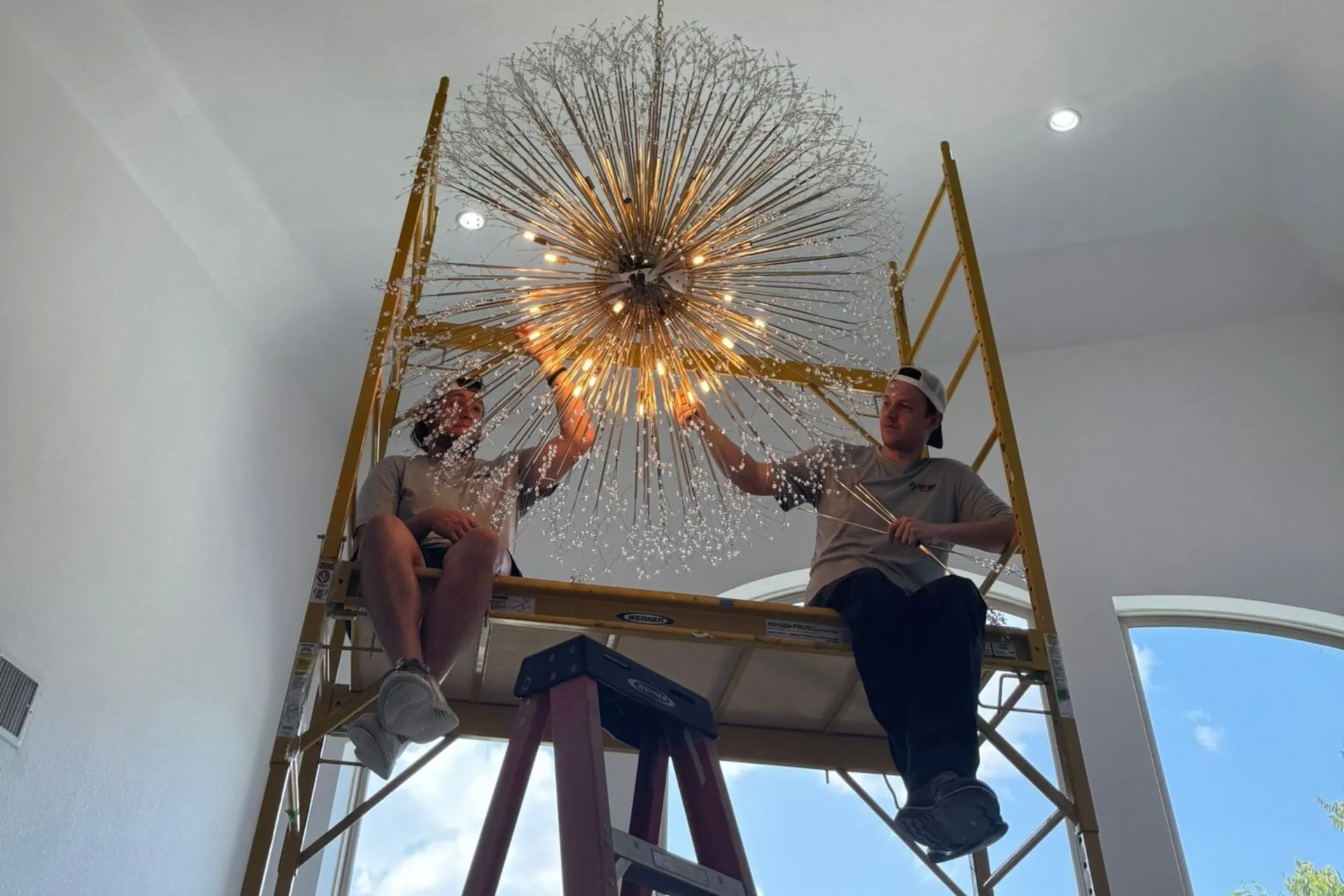 Tier One Electric technicians installing a large chandelier or ceiling light fixture in a high-ceiling interior space.
