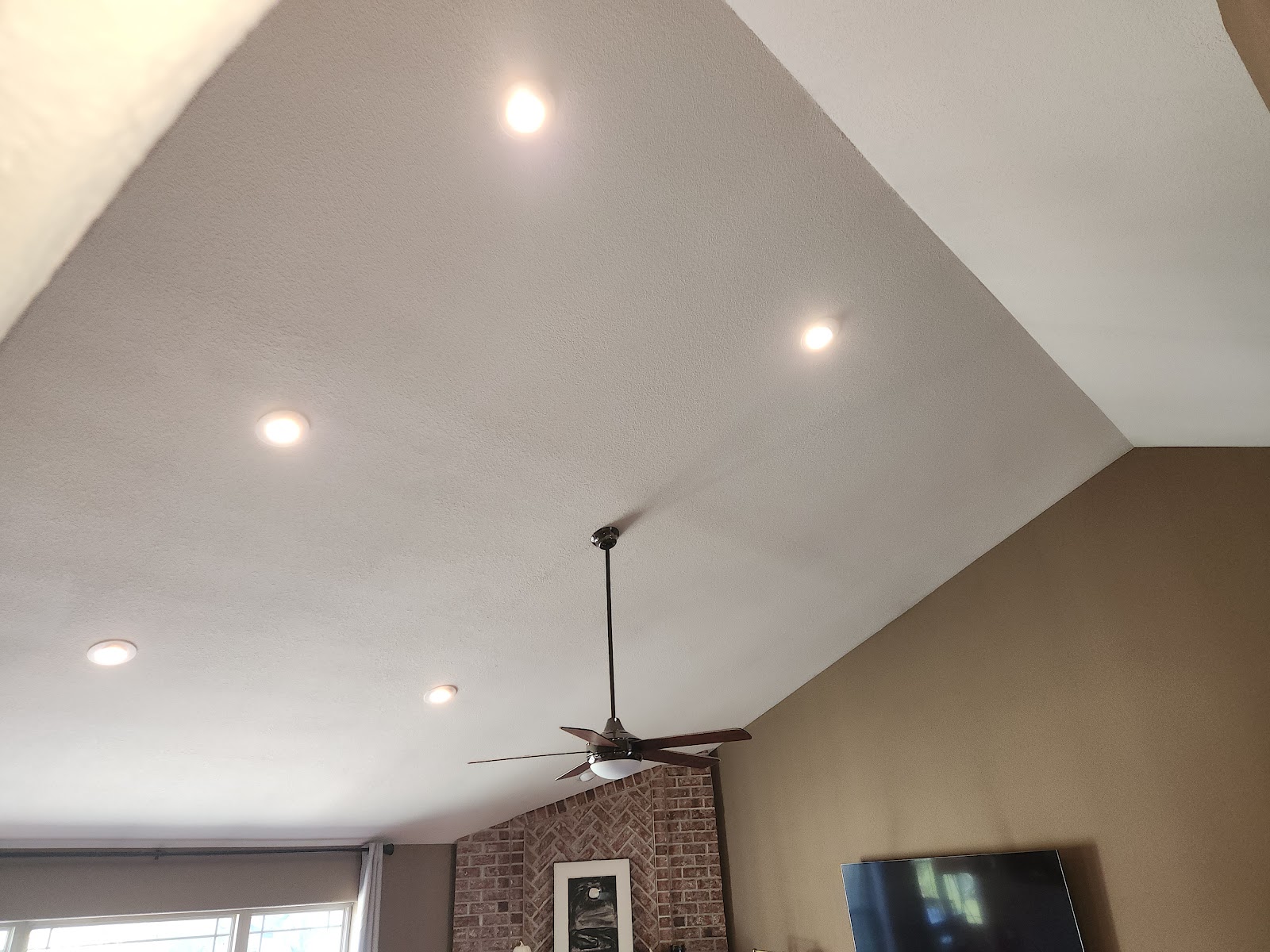 A vaulted ceiling with recessed lighting, a ceiling fan with wooden blades, and a section of brick wall with a framed picture is visible in this interior shot. The walls are painted a neutral tan color, and a large flat-screen television is mounted on the right side of the image.
