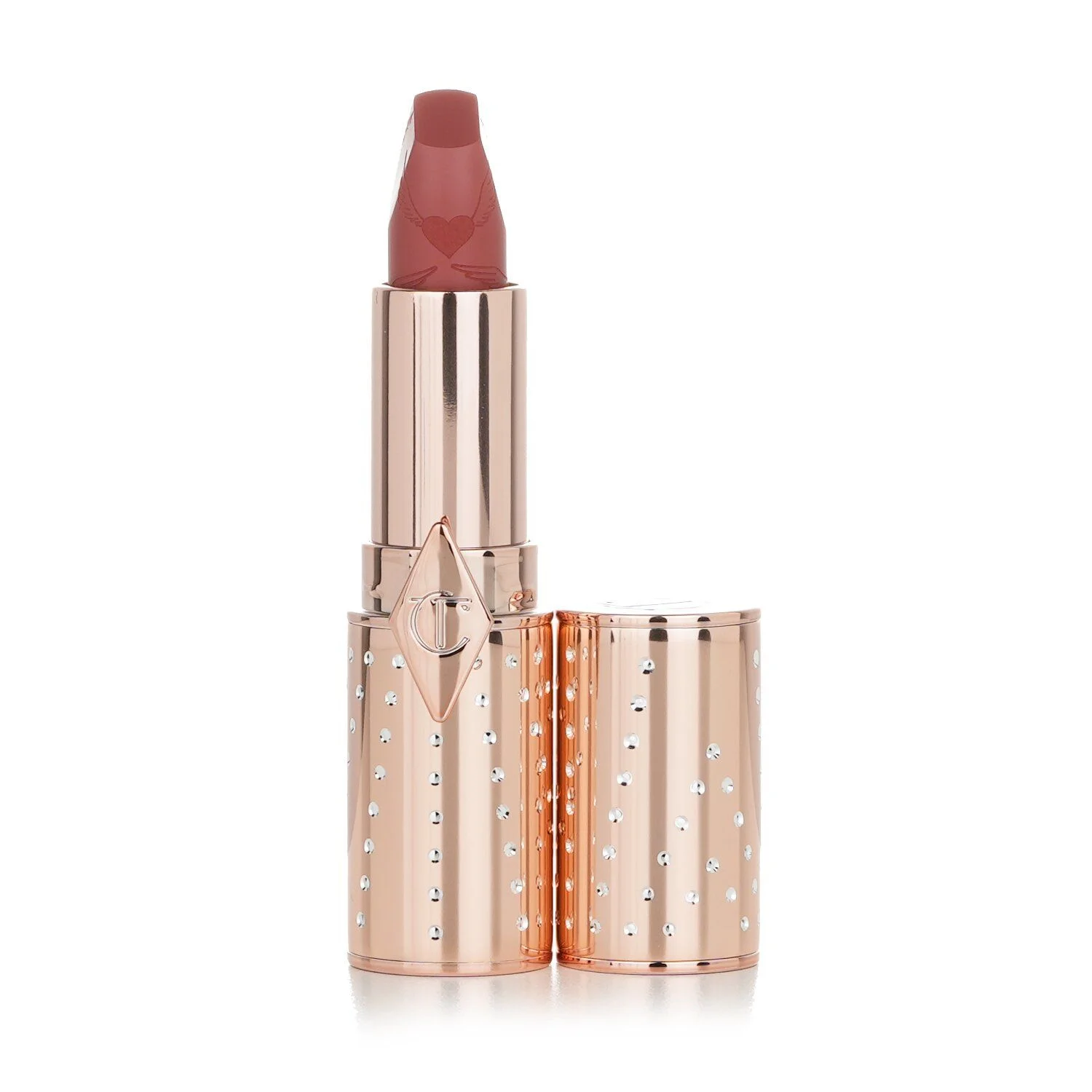 A close-up studio shot of a Charlotte Tilbury Matte Revolution lipstick in a rose gold case, against a white background. The lipstick bullet is a dusty rose shade and has a small engraved heart with wings design. The case is adorned with small, clear rhinestones.
