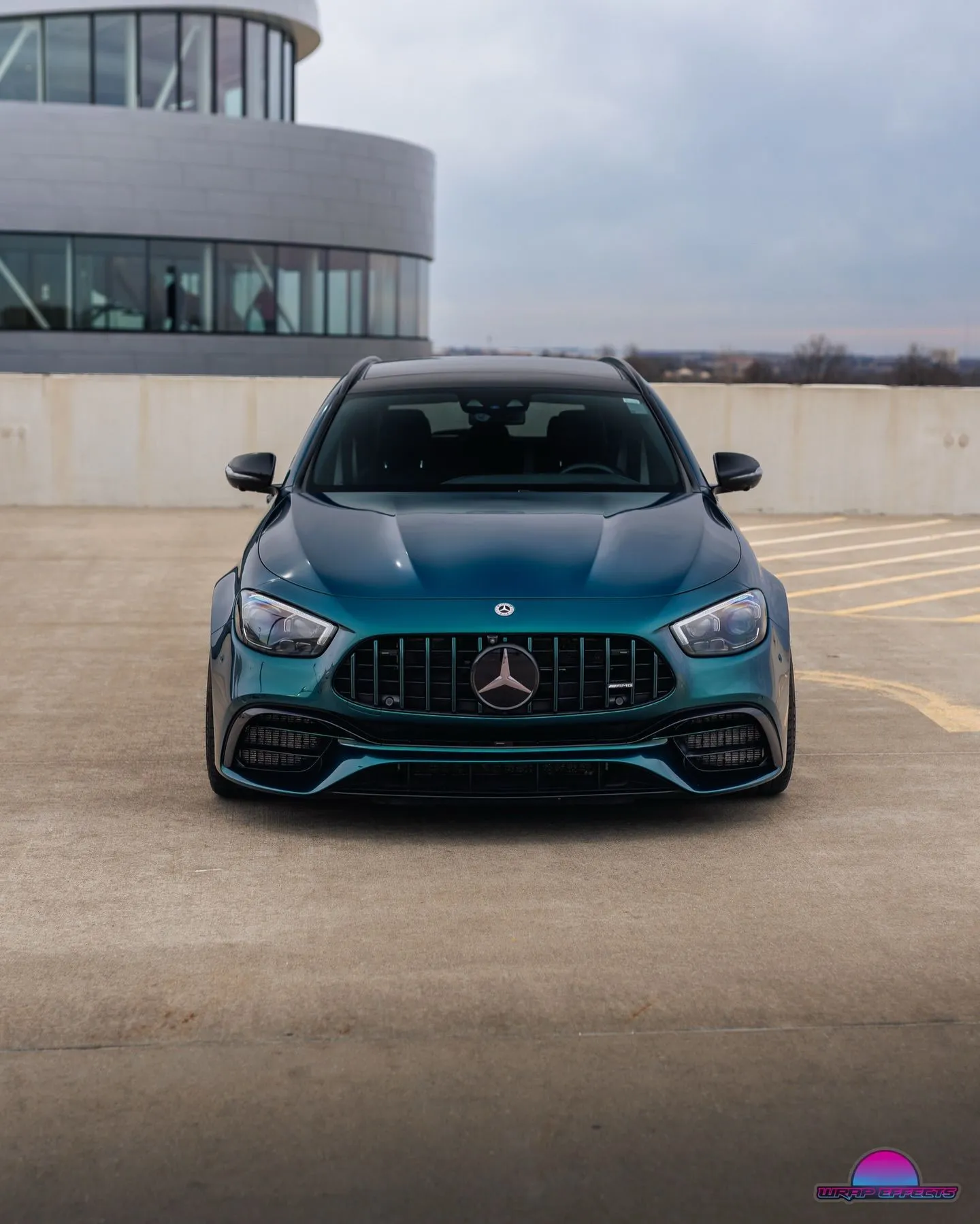 Front view of customized Mercedes-AMG performance wagon finished in metallic teal wrap, aggressive grille, sporty stance, and premium detailing photographed on rooftop parking structure with architectural backdrop.