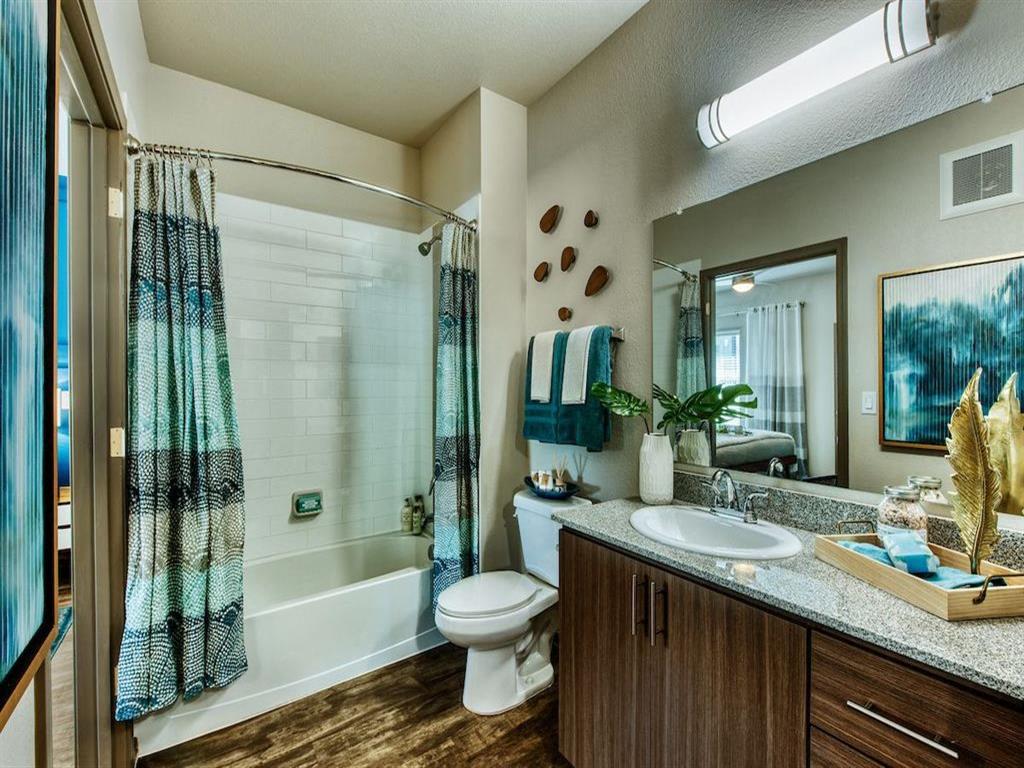 Apartment guest bathroom with white tub and blue shower curtain. Toilet can be seen next to a modern wood vanity and large mirror.