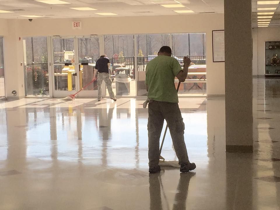 Janitorial staff mopping and cleaning large commercial lobby floor, maintaining shine and hygiene in high-traffic indoor space with natural light, glass doors, and visible reflections across polished surface.