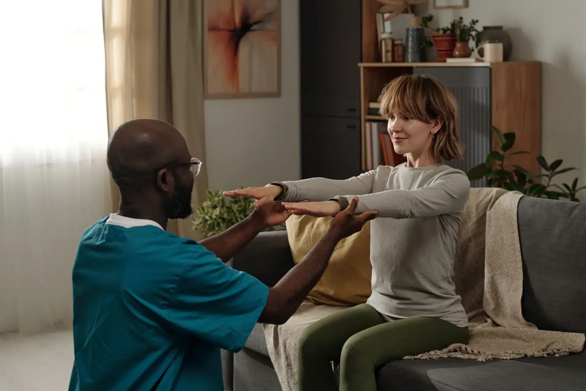 A male caregiver in a teal scrub top and a female patient in a gray long-sleeved shirt and green leggings are engaged in a physical therapy session. The caregiver is assisting the patient's arm movements. They are in a living room setting with a couch, a blanket draped over the side, and shelves filled with books and plants in the background. A framed piece of art hangs on the wall.