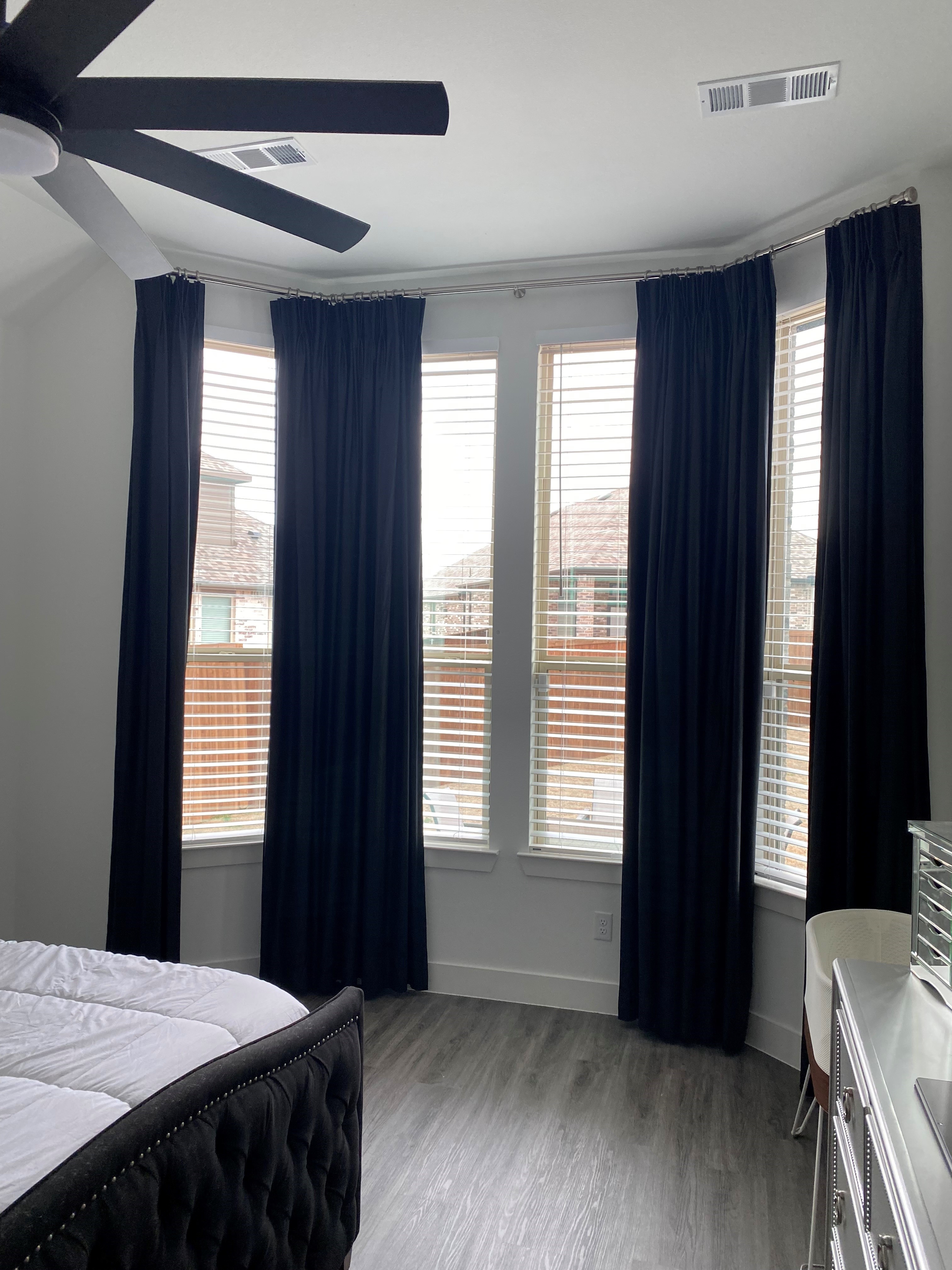 The team at Budget Blinds of Rockwall, Terrell, and Greenville absolutely love the clean, modern look of drapery paired with white faux wood blinds for bay windows in a master bedroom.