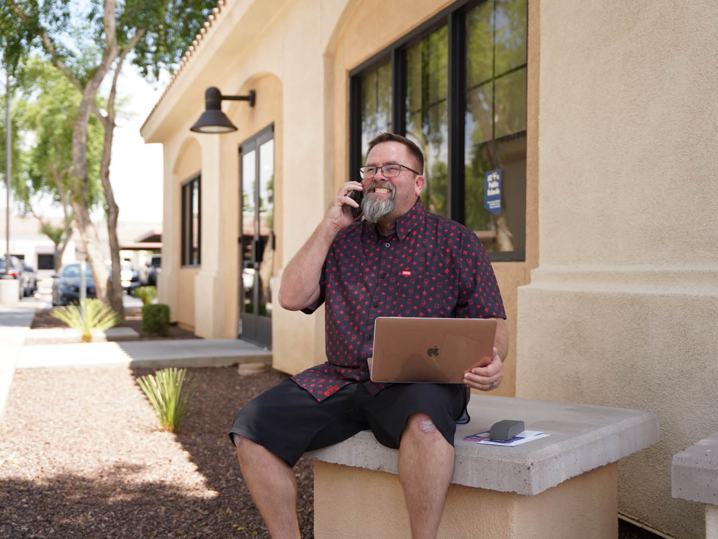 In this image, Mike Rux, a professional from Ciphers Digital Marketing, is captured outside the office in Gilbert, Arizona. He is sitting comfortably on a concrete bench, dressed in a casual patterned short-sleeve shirt and black shorts. With a laptop on his lap, Mike engages in a phone conversation, smiling and exuding a friendly and approachable demeanor. The building behind him features large windows and a light-colored exterior, and the surroundings include a well-maintained sidewalk and landscaping, enhancing the professional yet relaxed atmosphere of the location.