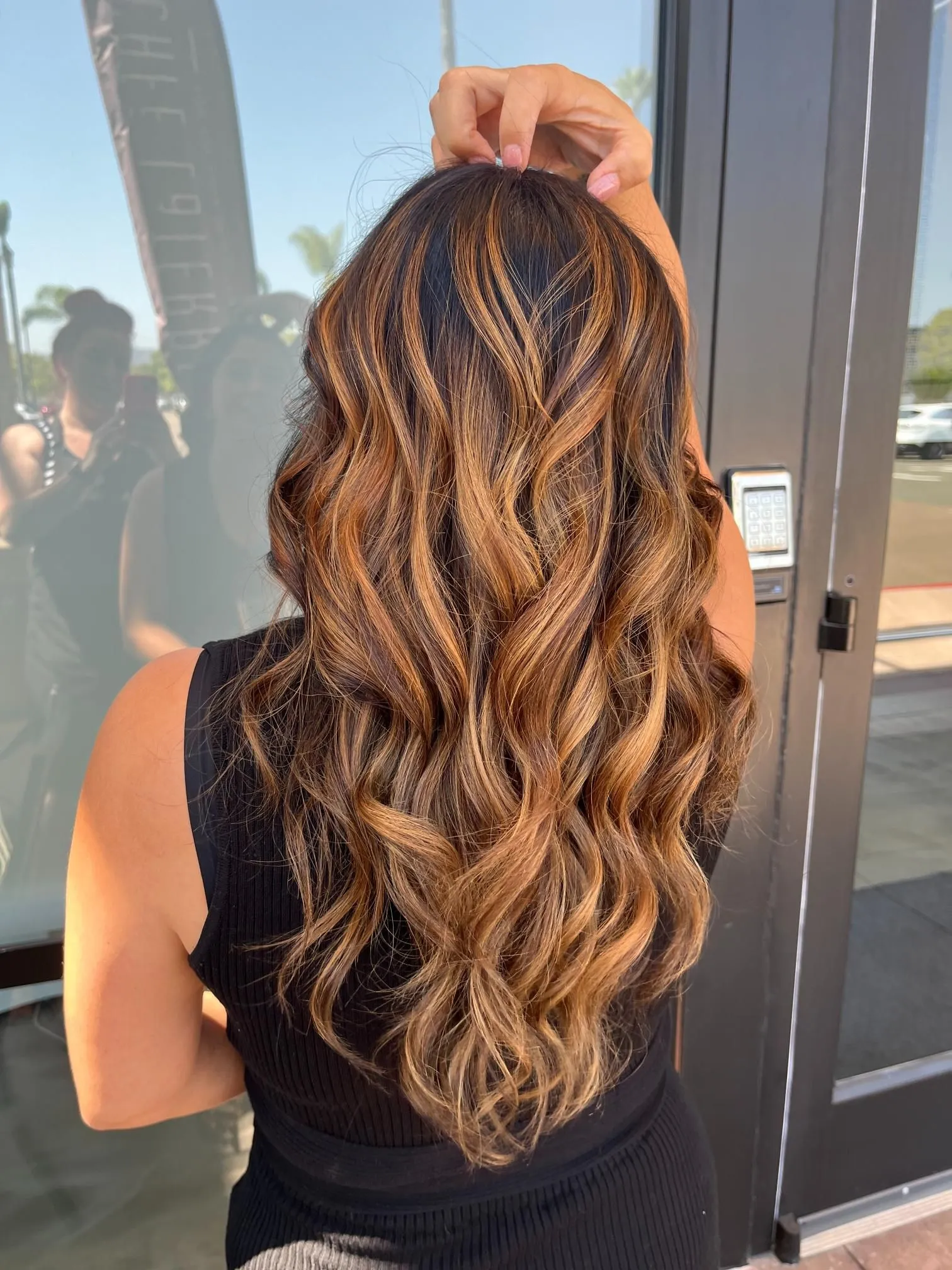 Long brunette hair with caramel balayage highlights styled in soft, flowing waves. The hairstyle showcases dimensional color blending and shine, photographed from the back to highlight professional salon coloring and styling.