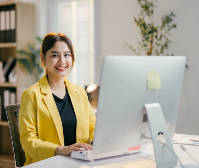 A woman wearing a yellow suit is sitting at a desk and working on a computer. 