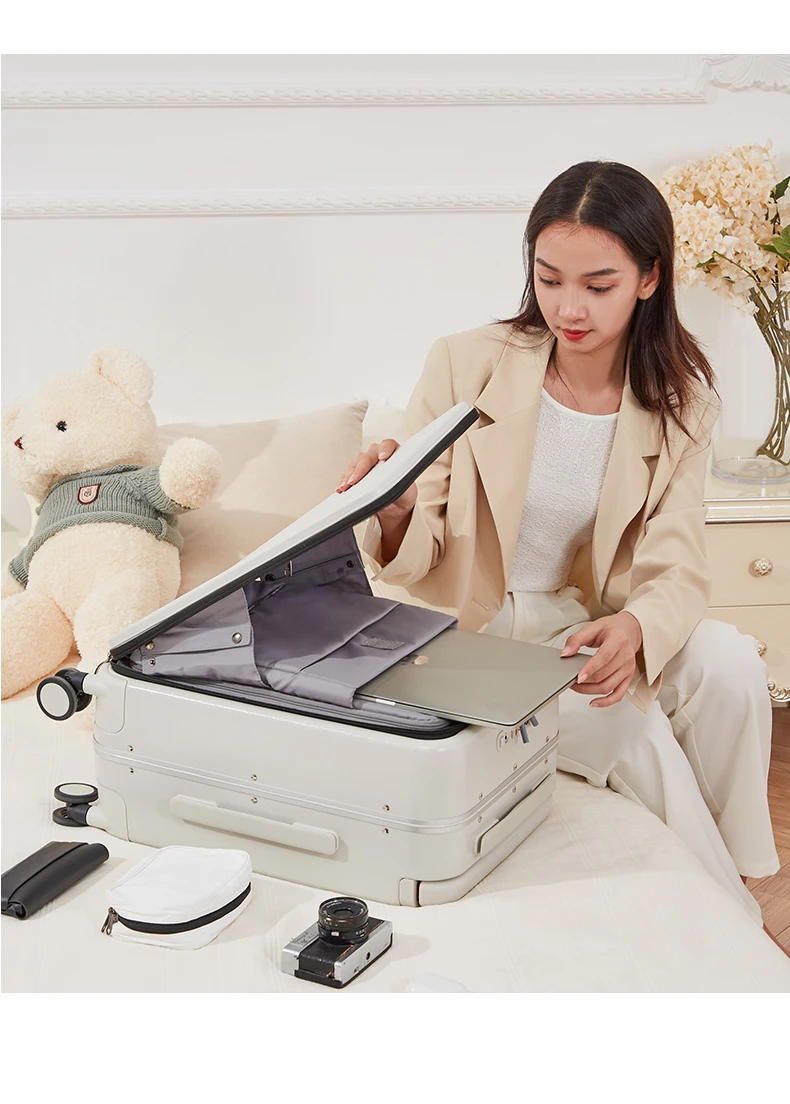 A young woman is packing a light-colored suitcase, placing a laptop inside. She is wearing a beige blazer over a white top and white pants. Beside the suitcase are a black clutch, a white toiletry bag, and a vintage camera. In the background, a large teddy bear sits on a bed, and a vase with flowers is on a nightstand.