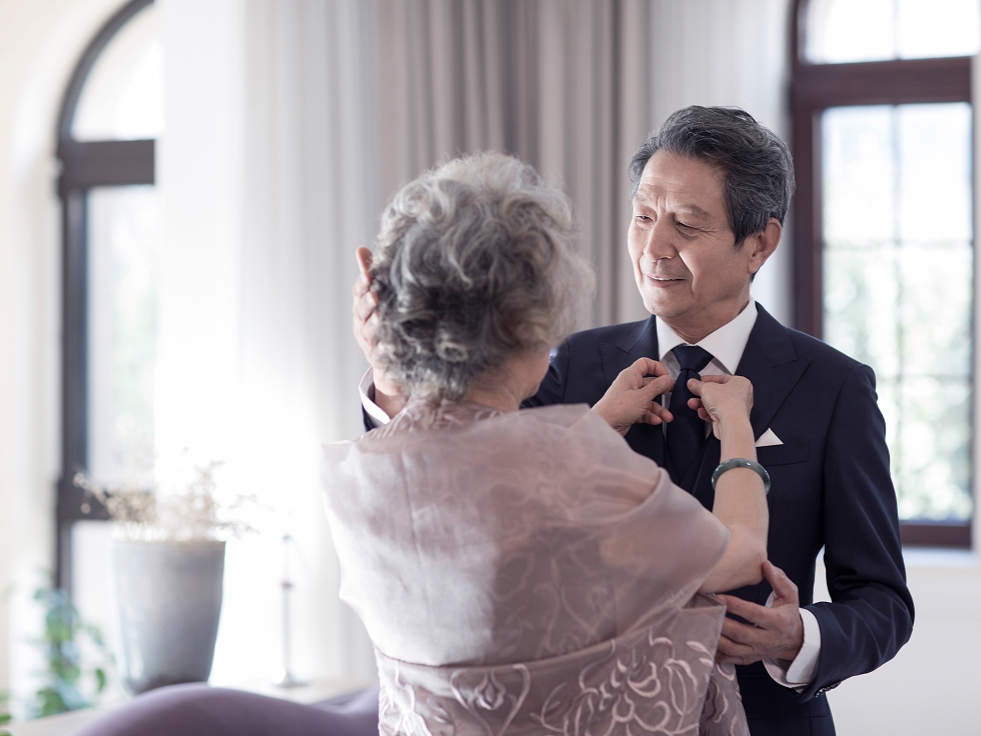 Elderly couple in a softly lit room; woman adjusts man's tie affectionately. Both are elegantly dressed, conveying warmth and love.