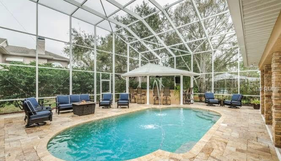 Screen-enclosed backyard swimming pool with clear blue water, stone paver decking, lounge seating, and a covered outdoor area.