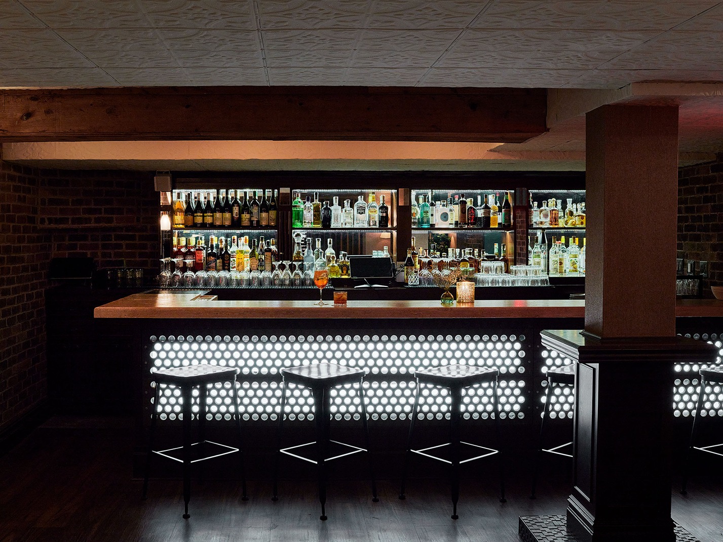 A dark, atmospheric bar area with a backlit, circular-patterned front. The back bar is brightly illuminated, highlighting rows of bottles and glassware in a moody setting.