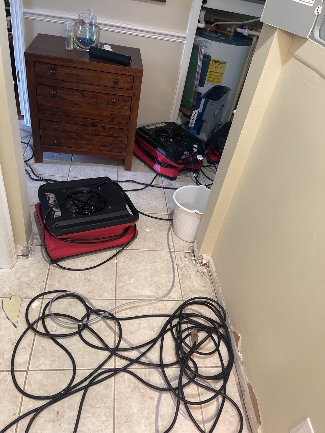 A scene of water damage remediation in progress, featuring industrial-style air movers and hoses connected to a water heater, alongside a wooden dresser. The flooring is tiled, and the walls show signs of recent damage and repair.