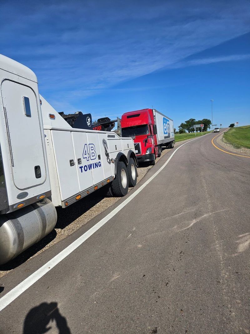 A large white 4B Towing commercial wrecker truck is parked along the shoulder of a curved asphalt highway, positioned to assist a red semi truck under a bright blue sky.