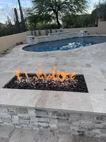 Scottsdale backyard with a built-in linear gas fire pit, black lava rock, and travertine coping, pool deck, and fire pit surround all matching. Sheer descent water feature visible in the background. Everything designed and built by Hacienda Outdoors, ROC #325952.