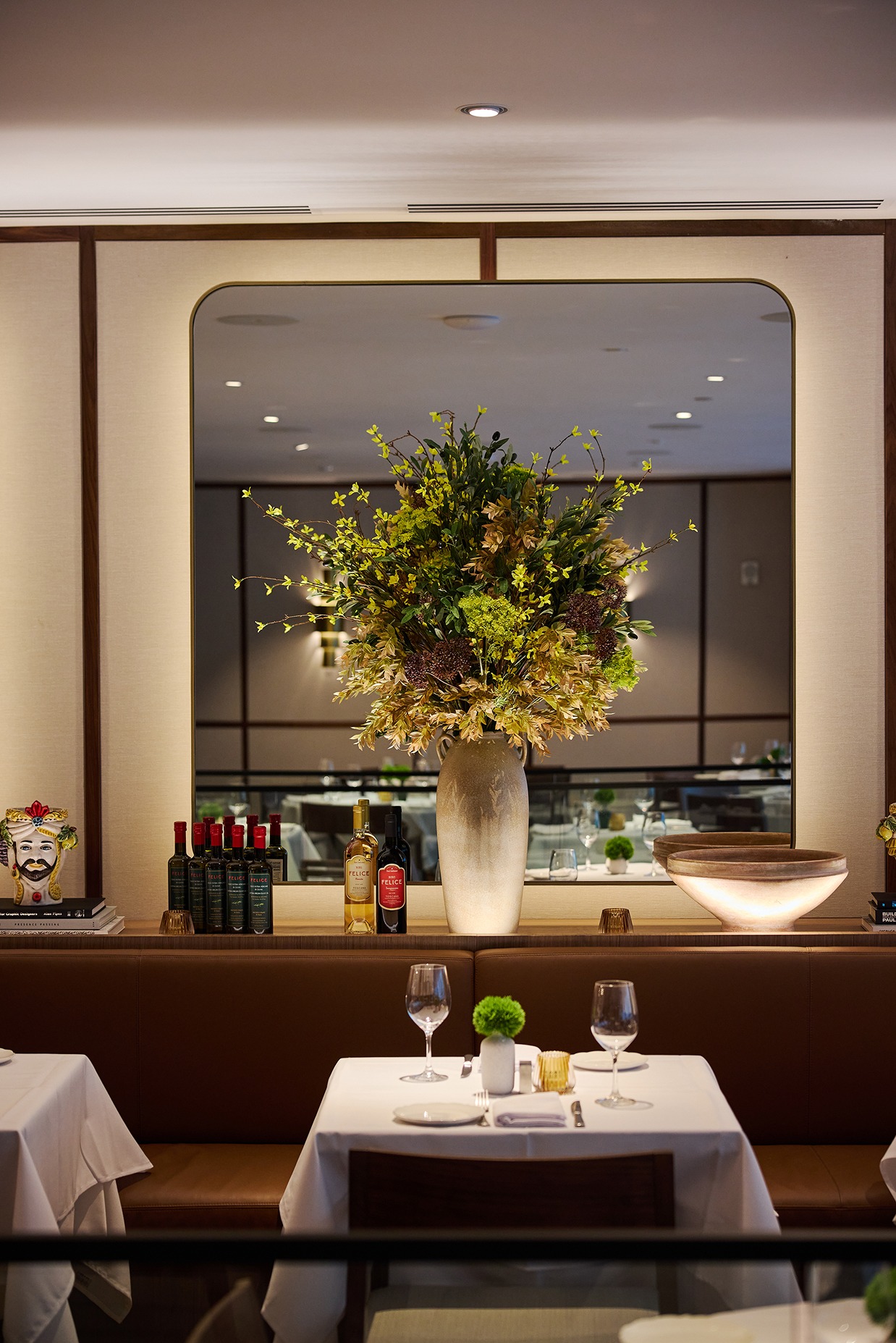 A white tablecloth dining setup with wine glasses and a lush green floral arrangement in front of a large mirror in a refined Italian eatery.