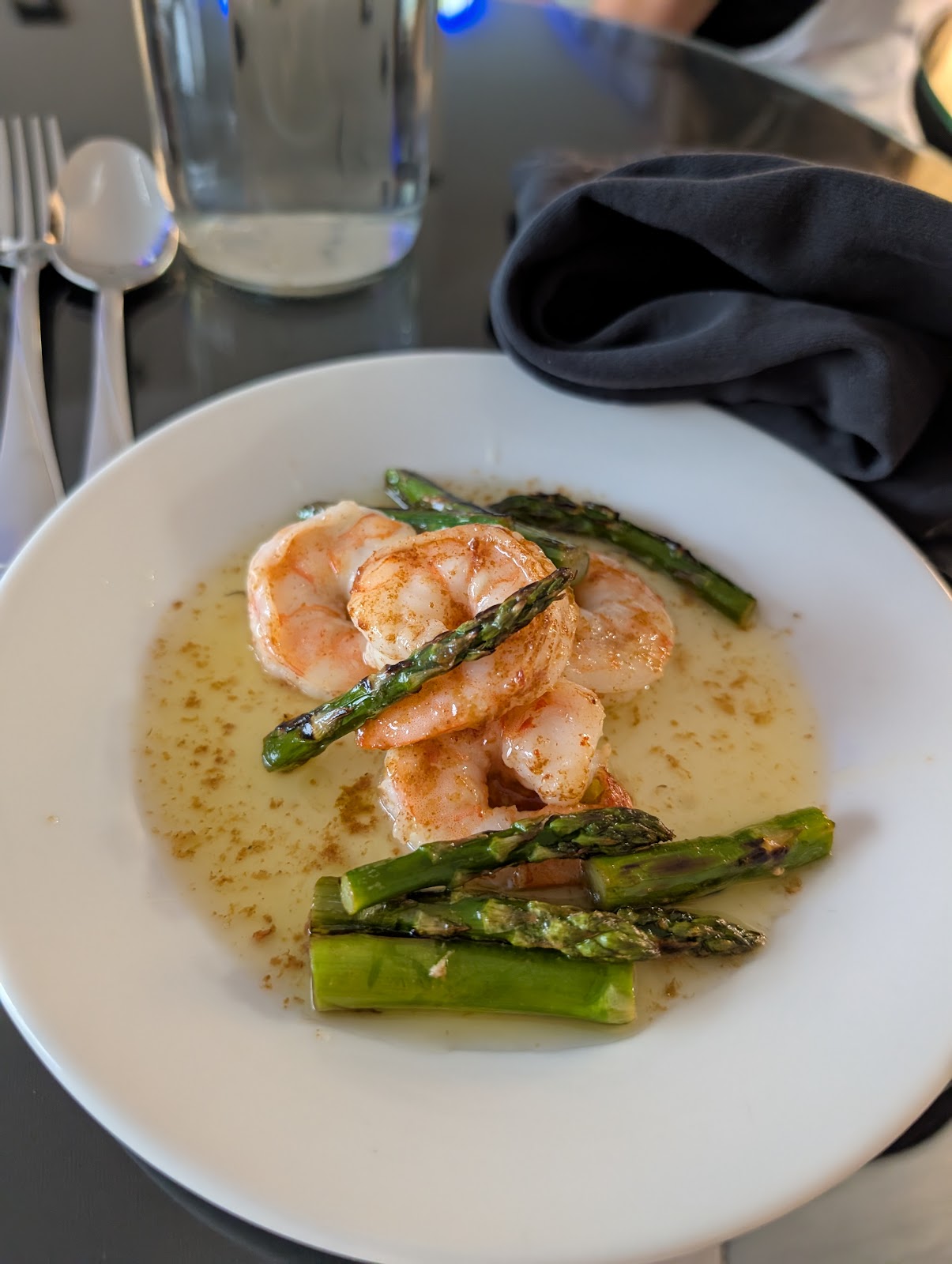 A close-up, high-angle shot of a white plate of shrimp and asparagus in a buttery sauce. The shrimp are large and pink, seasoned with paprika. The asparagus spears are grilled and vibrant green. The dish is served with silverware on the left and a dark napkin on the right. A glass of water is visible in the background.